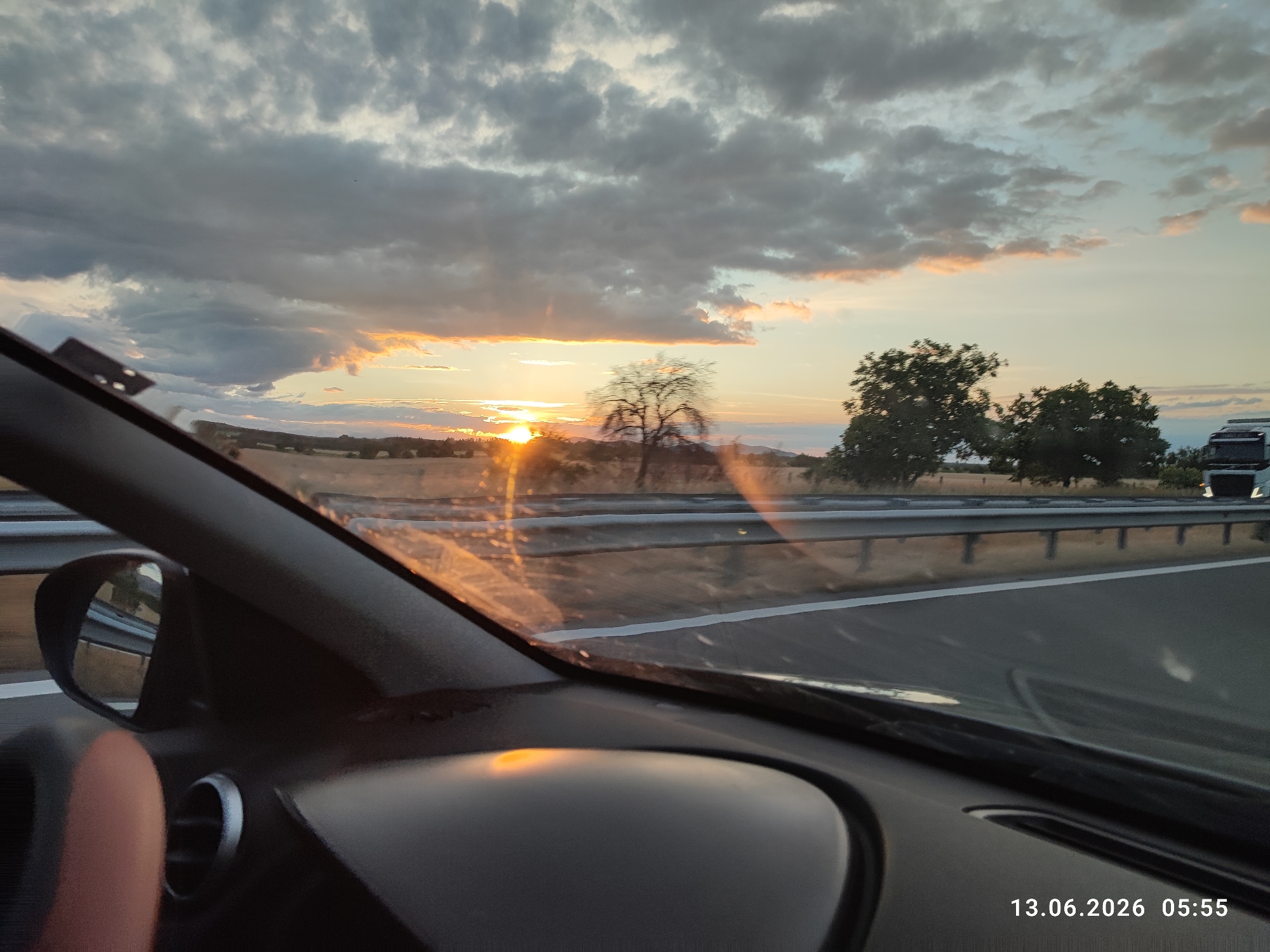 The image depicts a view from inside a vehicle, looking out through the windshield and side window. The time stamp indicates June 13, 2026, at 05:55. The scene appears to be a highway at sunrise, with the sun partially obscured by clouds and visible on the horizon. The sky is a mix of dark gray and lighter hues with hints of orange and pink from the sunrise. In the distance, there are trees and fields. A bare tree is prominent in the middle ground. The interior of the car is visible, including a side mirror, part of the dashboard, and a vent. The road itself is paved with lane markings. In the far distance on the right, a truck is partially visible. There are no people clearly visible within the frame. The location is identified as Shtarkovo, Bulgaria.