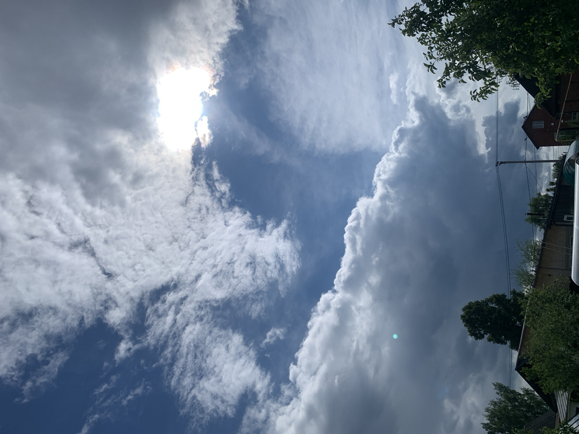 The image shows a sky with a mix of cumulus and cirrus clouds, some obscuring the sun. Patches of blue sky are visible. Tree branches and the roofs of buildings are visible at the top and right edge of the image. The location is likely Chaikovsky, Russia based on the prompt.