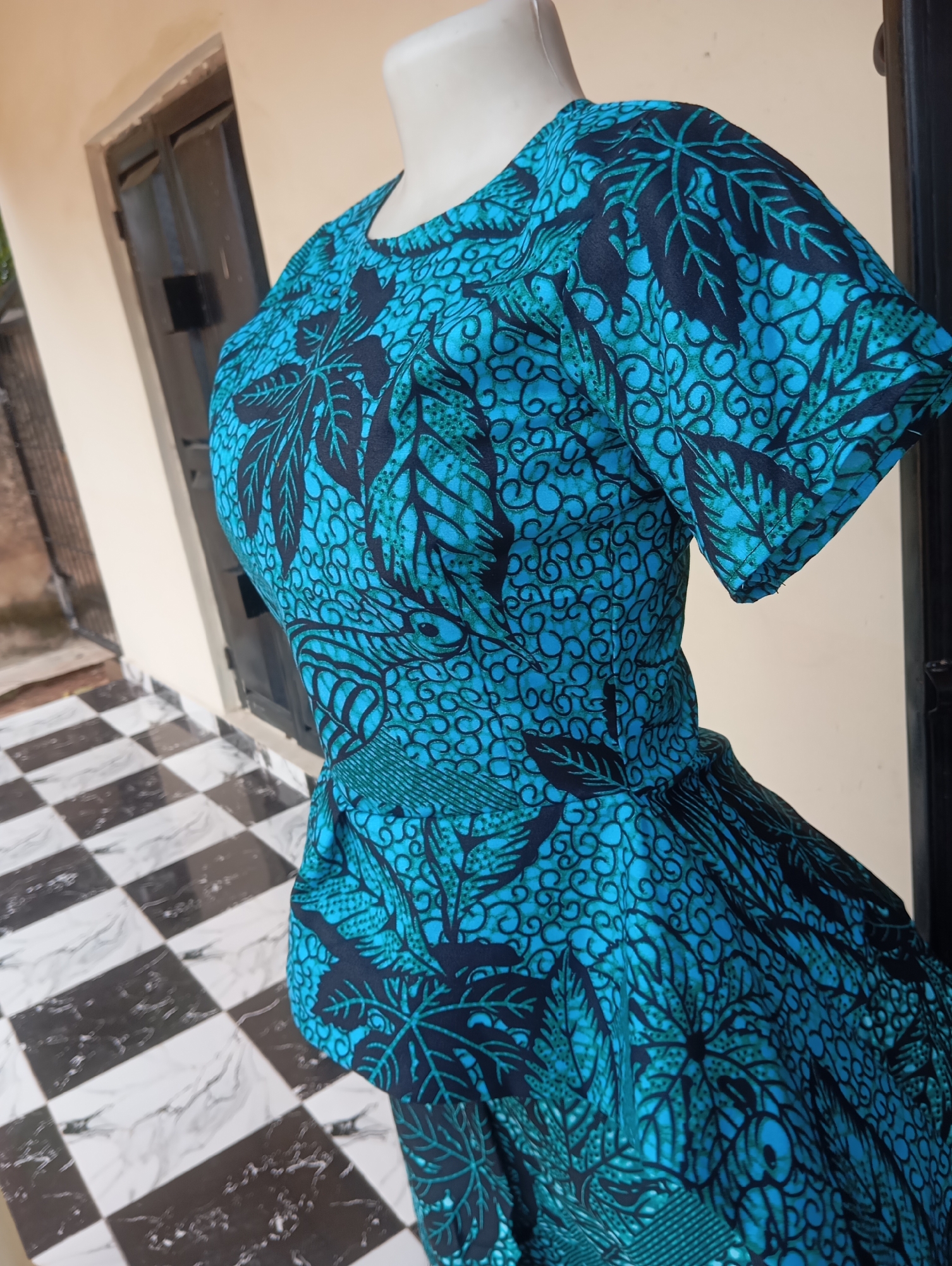 The image shows a close-up of a mannequin wearing a blue and black patterned dress. The dress has short sleeves and a flared skirt. The pattern on the dress features large leaf-like shapes and intricate swirling designs. The mannequin is positioned outdoors, in front of a building with a light-colored wall and a dark door or window. In the foreground, a black and white checkered tiled floor is visible. The lighting suggests it is daytime. The scene is likely located in Jalingo, Nigeria, given the context of the request.