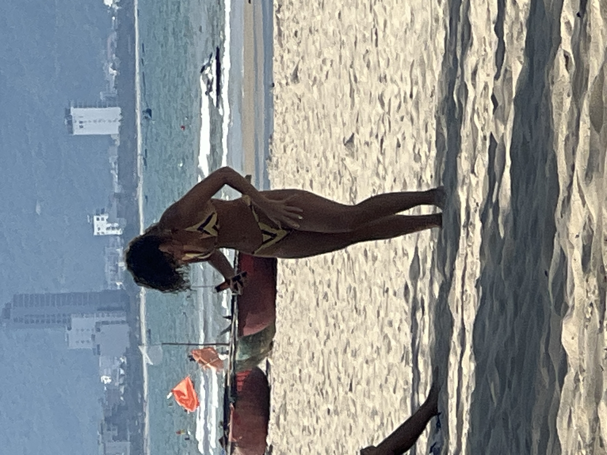 The image depicts a crowded beach scene in Đà Nẵng, Vietnam, with a woman in a bikini standing on the sand. She is looking down at her phone. In the background, the ocean is visible with a jet ski and boats. The shoreline is lined with fishing boats and orange flags. Buildings and mountains form the backdrop of the scene, indicating an urban coastal environment. The sand is light brown and appears to be densely packed.