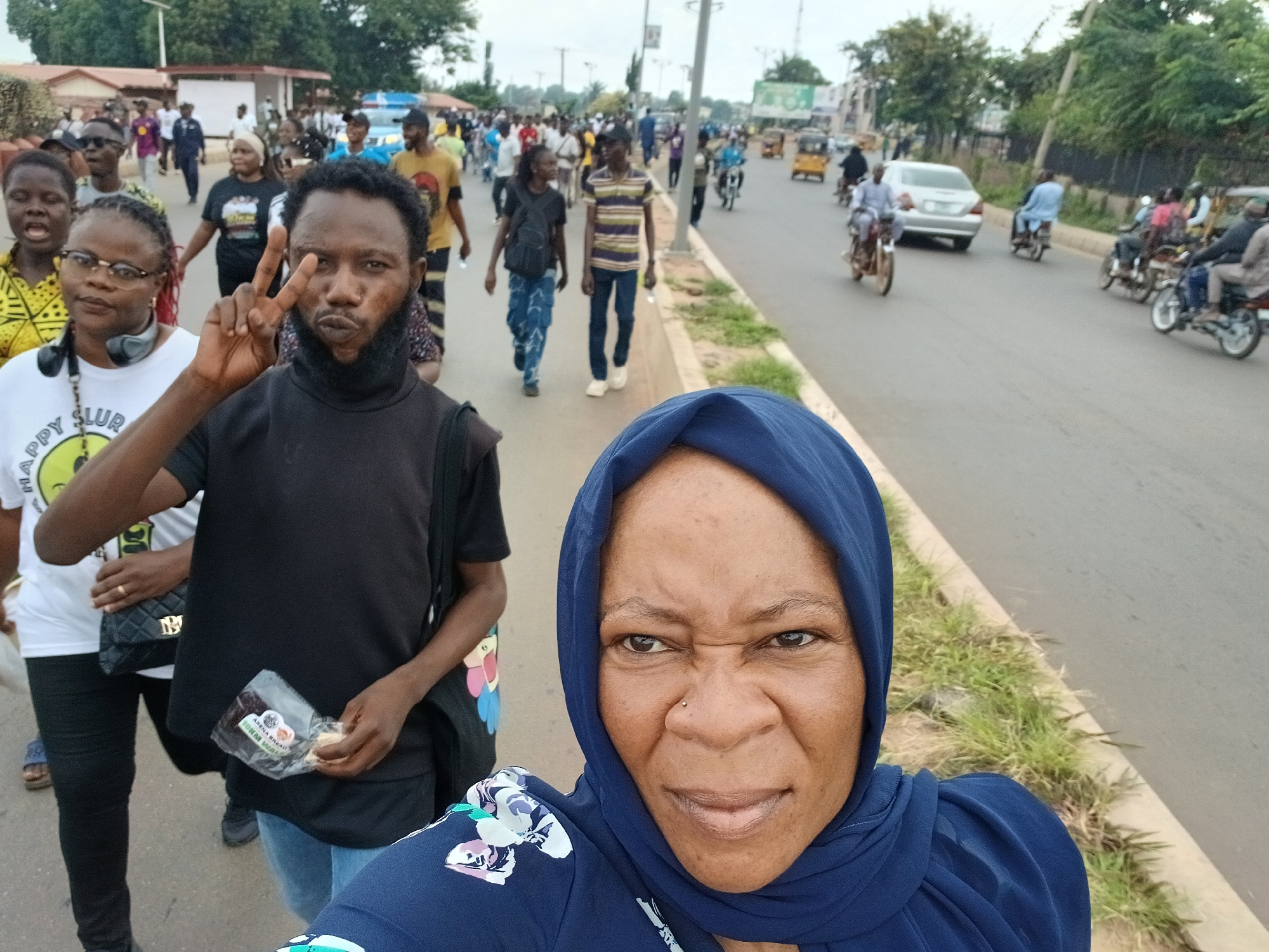 The image captures a street scene in Tudun Nupawa, Nigeria, with a group of people walking along a paved road. In the foreground, a woman wearing a blue hijab and patterned blue top is taking a selfie, her face angled towards the camera. To her left and slightly behind her, a man with a beard and a black top is holding up two fingers in a peace sign, and holding a small plastic bag with food items. Behind them, a woman with glasses and headphones around her neck, wearing a white t-shirt, walks alongside other pedestrians. Several individuals are visible in the background, including men and women walking, some carrying bags, and others on motorcycles. The road is lined with grassy verges and trees, and buildings can be seen in the distance. Traffic, including cars and motorcycles, is present on the road, indicating a moderately busy urban environment. The sky is overcast.