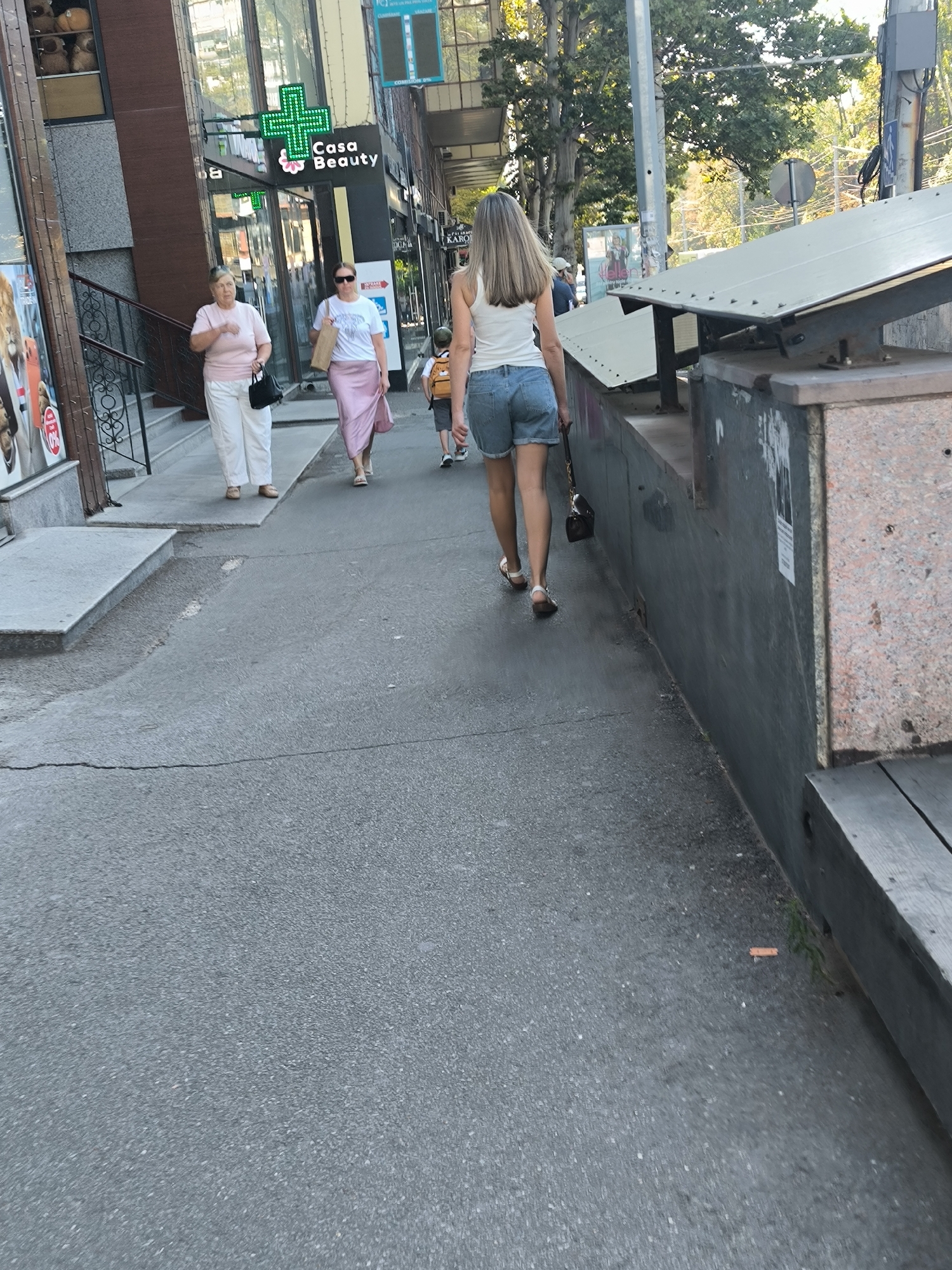 The image shows a street scene in Chișinău, Moldova, with several people walking on a sidewalk. In the foreground, the asphalt is cracked and worn. To the left, a woman in a pink shirt and white pants walks away from the camera, carrying a black purse. Next to her, another woman in a white t-shirt and a long pink skirt also walks away, carrying a brown paper bag and wearing sunglasses. Further down the sidewalk, a young person in denim shorts and a white tank top walks away from the camera, holding a dark purse. The background features buildings, including a storefront with a green cross sign and the words "Casa Beauty". Trees and a utility pole are also visible. A cigarette butt is lying on the ground near a wooden bench. The lighting suggests it is daytime, possibly late afternoon, with shadows cast by the buildings and trees.