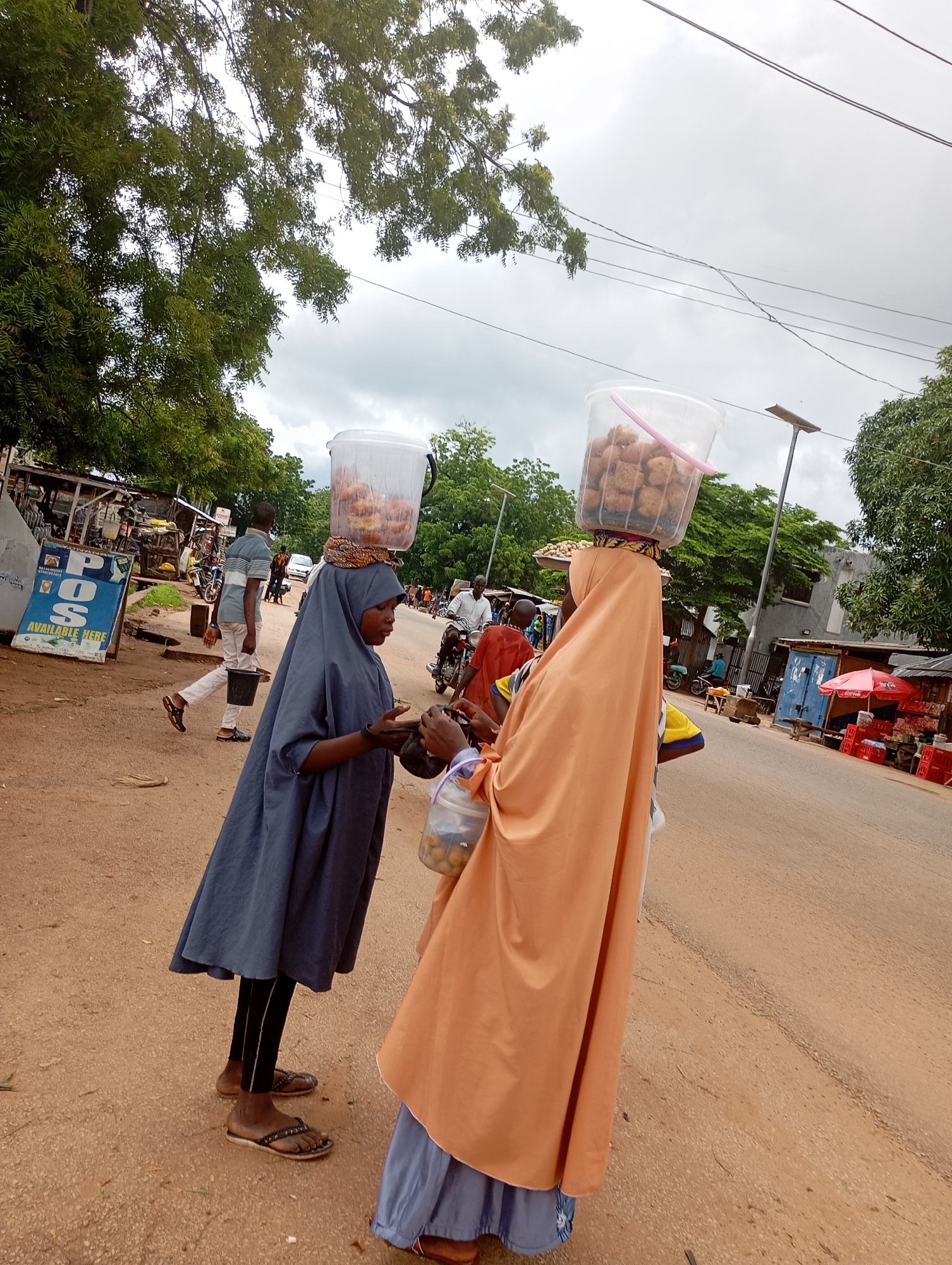 In the city of Shendam, Nigeria, two women are seen standing on a dusty street. The woman on the left, dressed in a dark blue hijab and covering, balances a large clear plastic bucket filled with food items on her head. She appears to be interacting with the woman on the right, who is draped in a vibrant orange hijab and covering, also carrying a similar bucket of food. Both women are wearing traditional Islamic attire. In the background, other people, vehicles including motorcycles, and various stalls and shops line the street, indicating a bustling marketplace or commercial area. The environment suggests a warm, possibly humid climate with cloudy skies overhead. A sign with the text "POS AVAILABLE HERE" is visible on the left side of the image, suggesting the presence of a financial service point. The ground is covered in red dirt, typical of many regions in Nigeria.