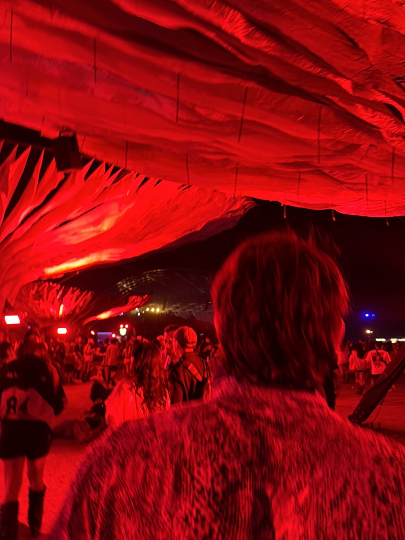 The image shows a crowd of people at an event in Indio, California. A red-toned, tent-like canopy covers the area. A person in a patterned shirt is in the foreground, with their back to the camera. Other people are visible in the distance, along with blurred objects and lights.