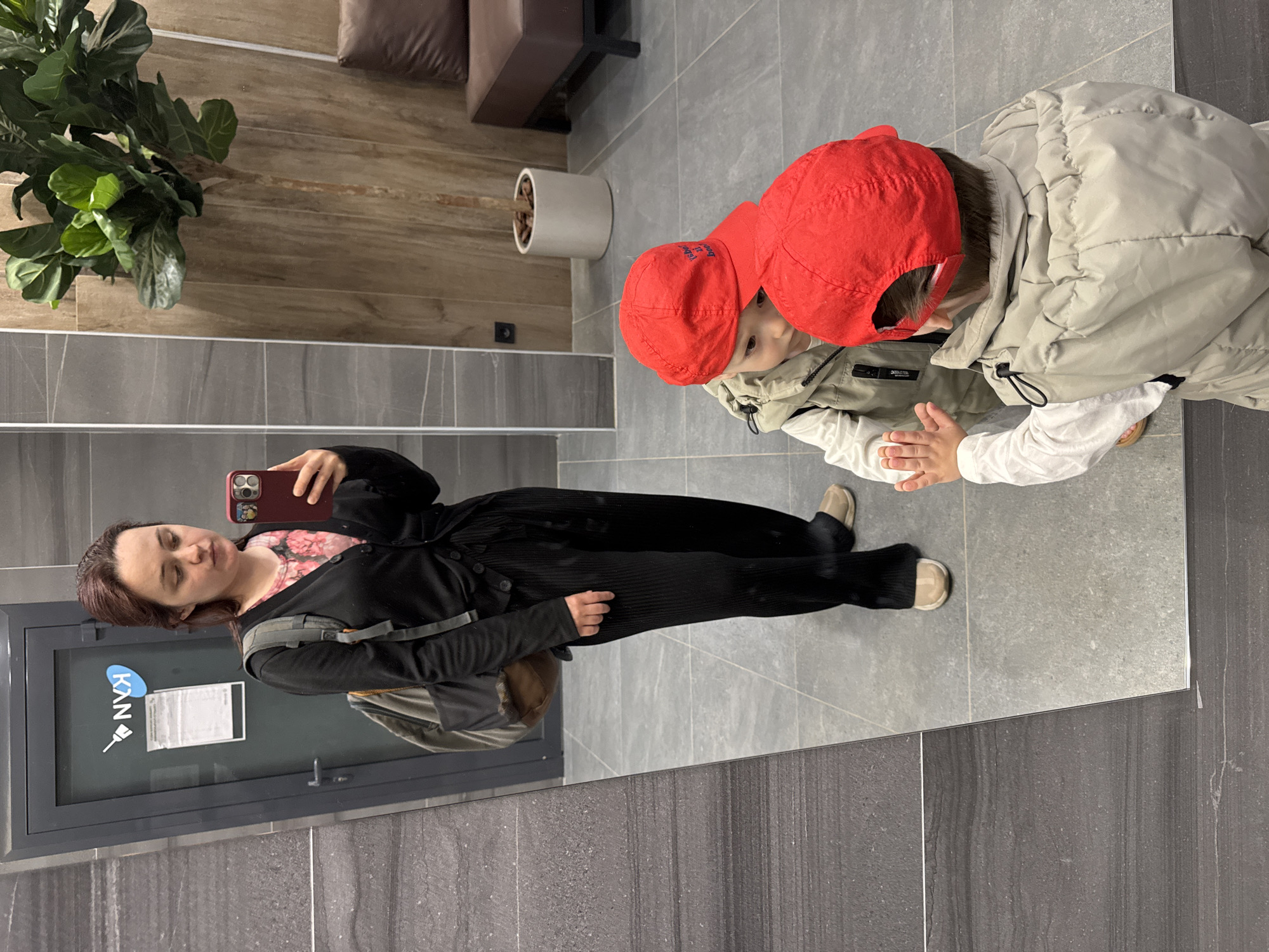 The image captures an indoor scene, likely a modern lobby or hallway in Izhevsk, Russia. A woman is seen in a mirror reflection, taking a selfie with her smartphone, which has a red case. She has dark hair, wears a black cardigan over a pink floral top, black ribbed pants, and beige slip-on shoes, with a dark backpack on one shoulder. Her expression is neutral.
To the right of the reflection, a young child is visible, facing towards the mirror. The child wears a red baseball cap with indistinct text (possibly "USO 21"), a light beige padded winter jacket, and a white long-sleeved shirt. Their hands are slightly raised, and they appear to be looking towards the woman or the phone.
The setting includes light gray tiled flooring and a darker gray, textured wall on the left. Above the tiled section, the wall is clad in light brown wooden panels. A large potted plant with dark green leaves, a section of a brown sofa cushion, and a white cylindrical planter are visible in the background.
On a door or panel reflected in the mirror, there is a light blue sticker displaying the text "KAN" alongside a small wrench symbol. A white paper document is taped below the sticker, but its text is illegible. The overall scene depicts a casual moment captured in a well-lit, contemporary interior.