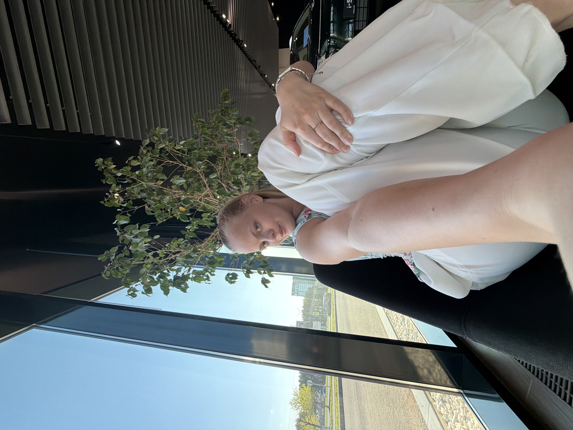 A woman sits in a black chair inside a building with large windows, likely a showroom, with a tree next to the window. She wears white trousers and a floral top, hugging her legs. Her left hand, adorned with a silver bracelet and a ring, rests on her knees. Outside the window, a cityscape is visible under a bright blue sky. The location appears to be Izhevsk, Russia, based on the architectural style of the visible buildings and the landscaping. The building's interior has a modern design with a dark ceiling and distinctive vertical elements.