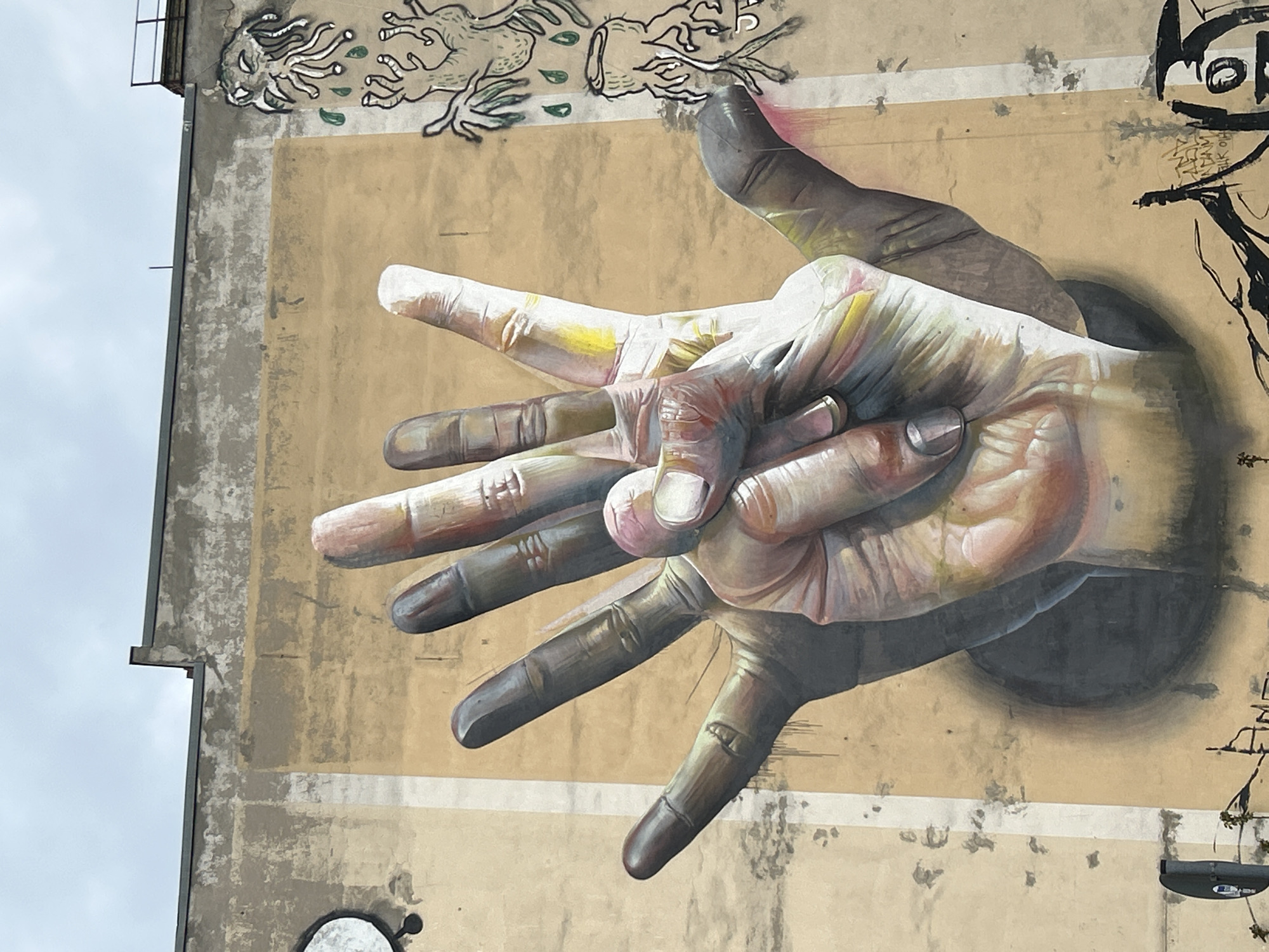 The image shows a mural painted on the side of a building in Berlin, Germany. The main subject of the mural is a pair of hands depicted in a photorealistic style. One hand is white or light-skinned, while the other hand is black or dark-skinned, appearing to reach out from a hole or opening in the wall. The hands are intertwined, with their fingers overlapping. The skin tones of the hands have been carefully rendered with variations in color and shading to create a lifelike appearance. The background of the mural is a beige or tan color, and there are other stylized designs or graffiti elements painted around the hands, including creatures or abstract shapes. The building itself appears to be older, with visible signs of wear and tear on the facade.