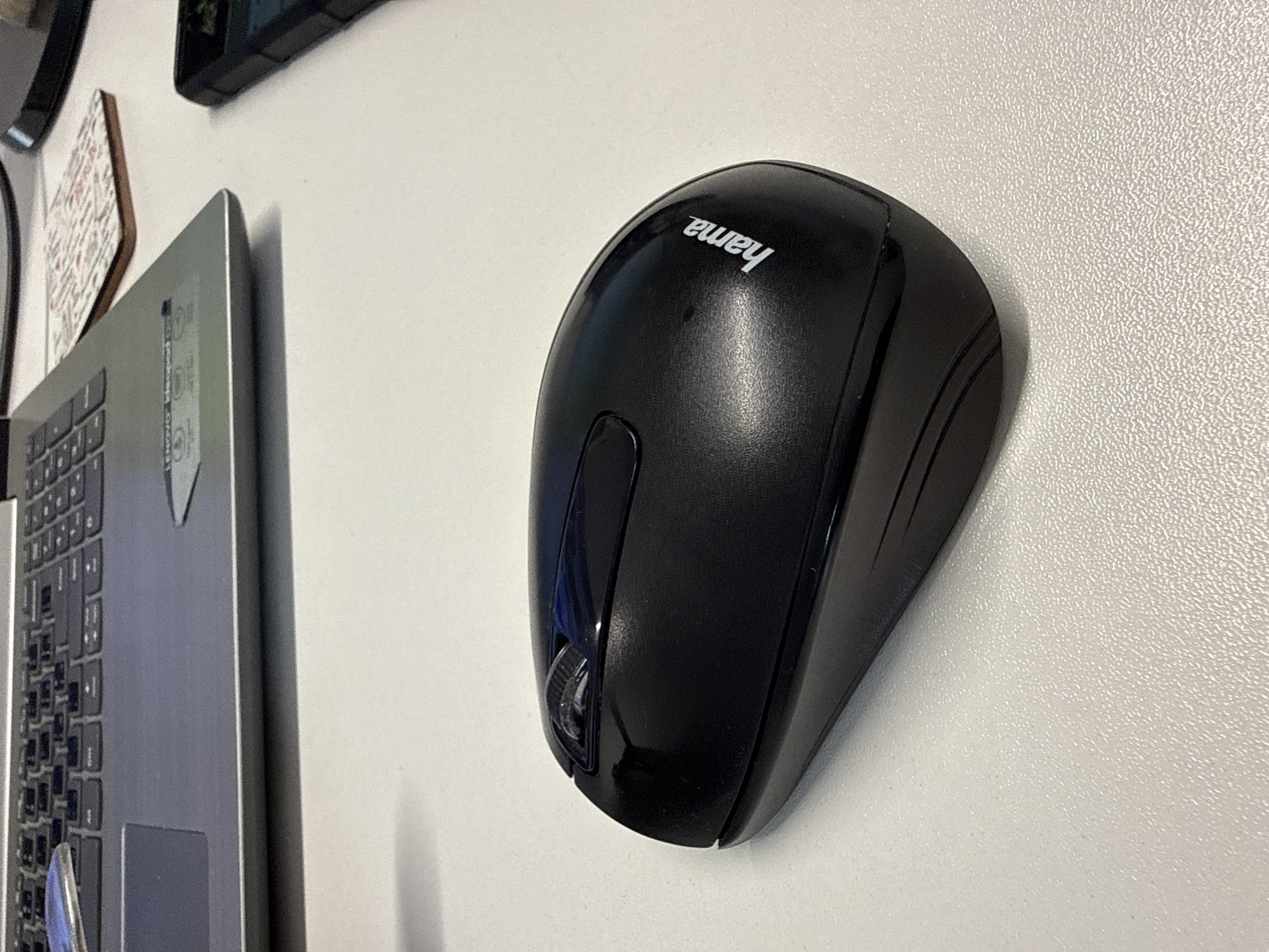 A black wireless computer mouse with the brand name "hama" printed in white is placed on a white textured surface. To the left and slightly behind the mouse, a silver laptop is partially visible, with its keyboard and palm rest in focus. The laptop appears to be open. In the background, a corner of a black smartphone is visible on the right side of the frame. The scene is indoors, likely on a desk, and there is no indication of the city of Łódź, Poland.