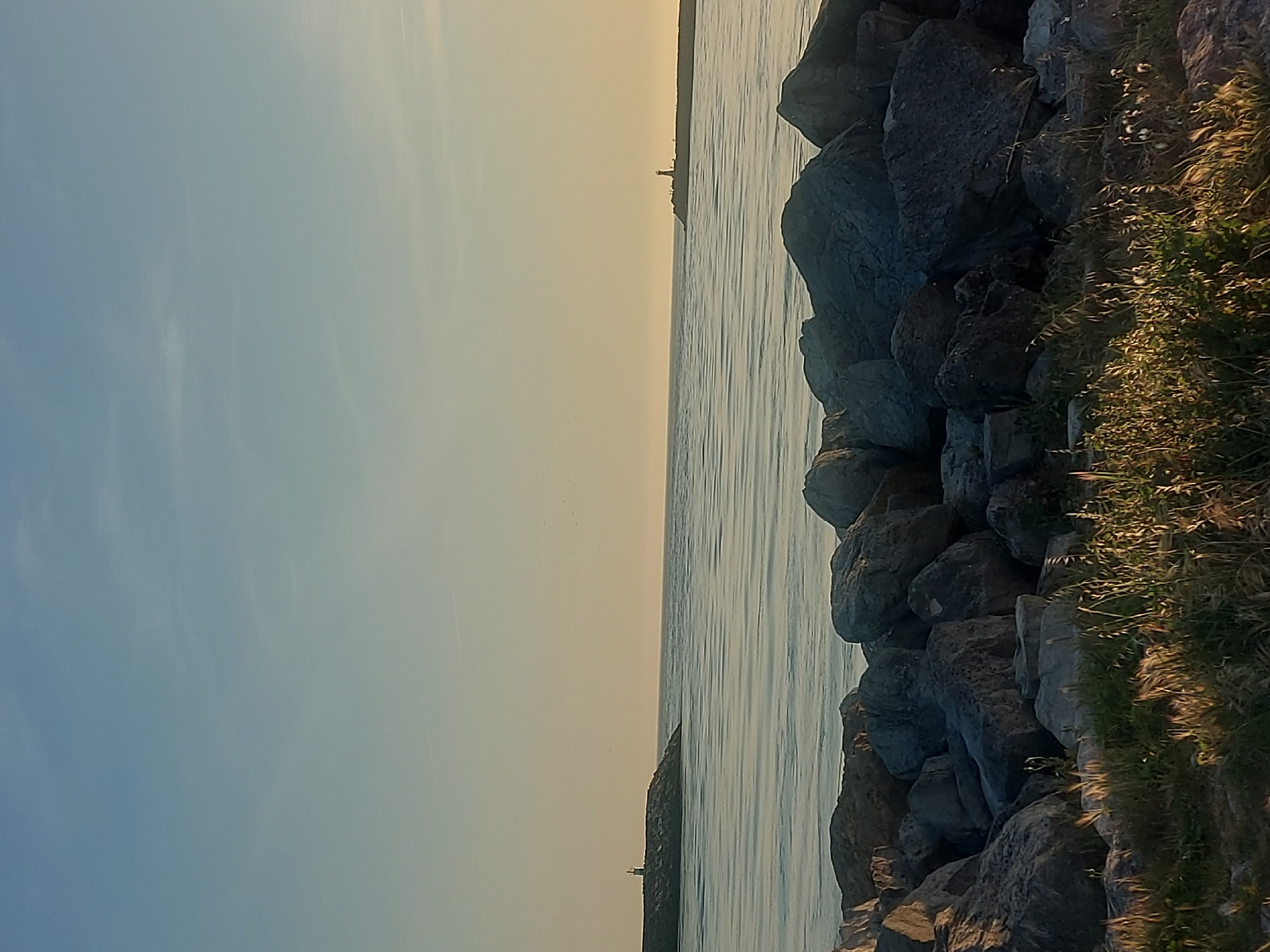 The image depicts a coastal scene in Figueira da Foz, Portugal. On the right side are large grey rocks forming a barrier or breakwater, with some vegetation growing among them. The water extends towards the center, reflecting the sky. A faint structure resembling a lighthouse is visible in the distance. The sky on the left ranges from blue to yellow, suggesting either sunrise or sunset. There are no people visible in the image.