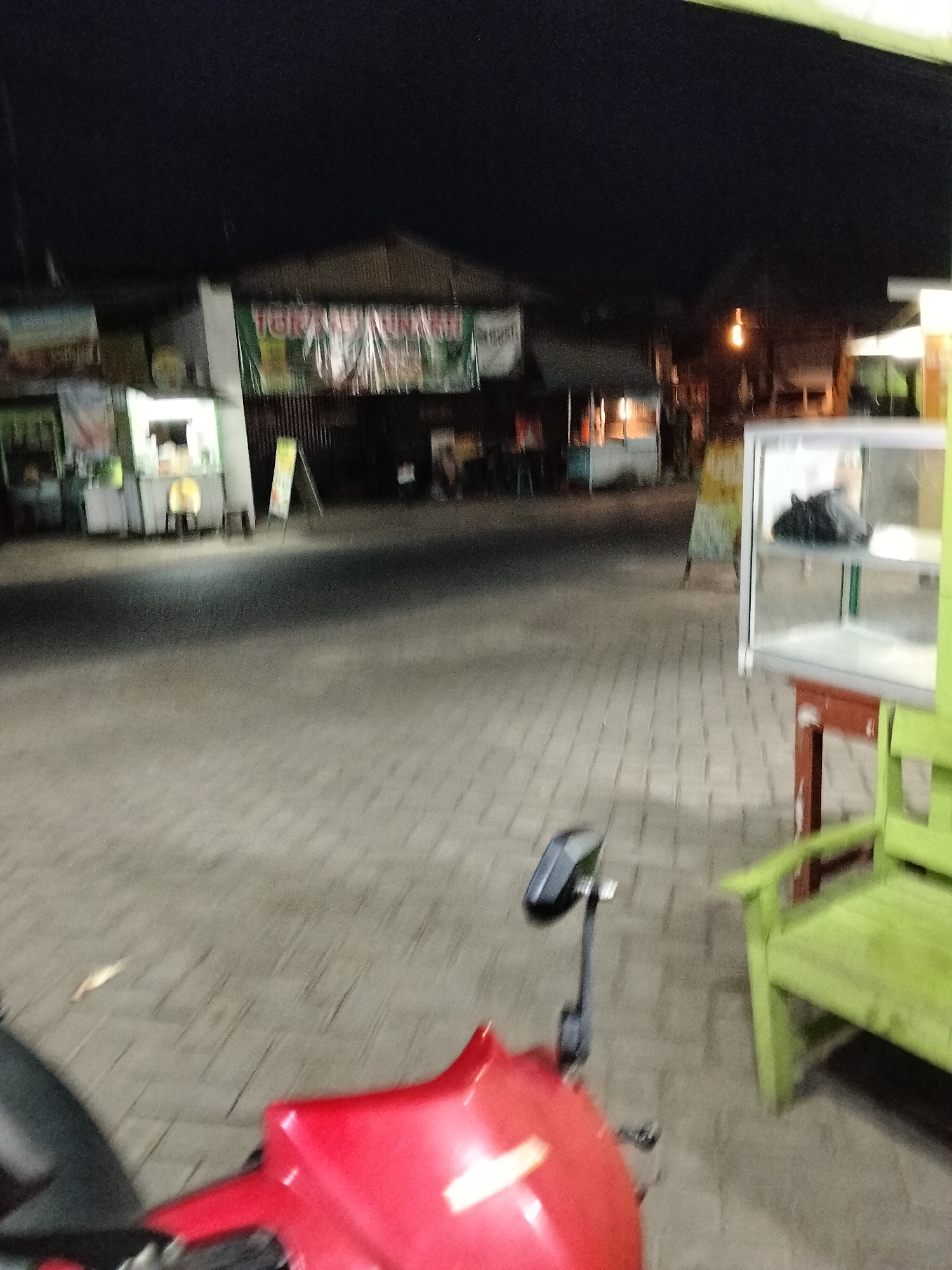 The image depicts an outdoor scene at night, likely in Gempolan, Indonesia, showing a street or walkway with several stalls and a motorcycle in the foreground. The area is paved with interlocking bricks.
On the left side of the image, there are several food stalls or kiosks. One stall is brightly lit, with a white facade and displays items inside. Another stall to its left has a banner with text, and other stalls are visible further back. A signpost is also present in front of one of the stalls.
To the right, a glass display cabinet is positioned on a red and green wooden table. A green wooden bench is situated next to the cabinet. The background shows more stalls and buildings, dimly lit, with some signs visible. A warm-toned light hangs from the ceiling of one of the stalls.
In the foreground, the red body of a motorcycle is prominent, with a black rearview mirror attached to its handlebar. The image is slightly blurred, suggesting movement or low light conditions.