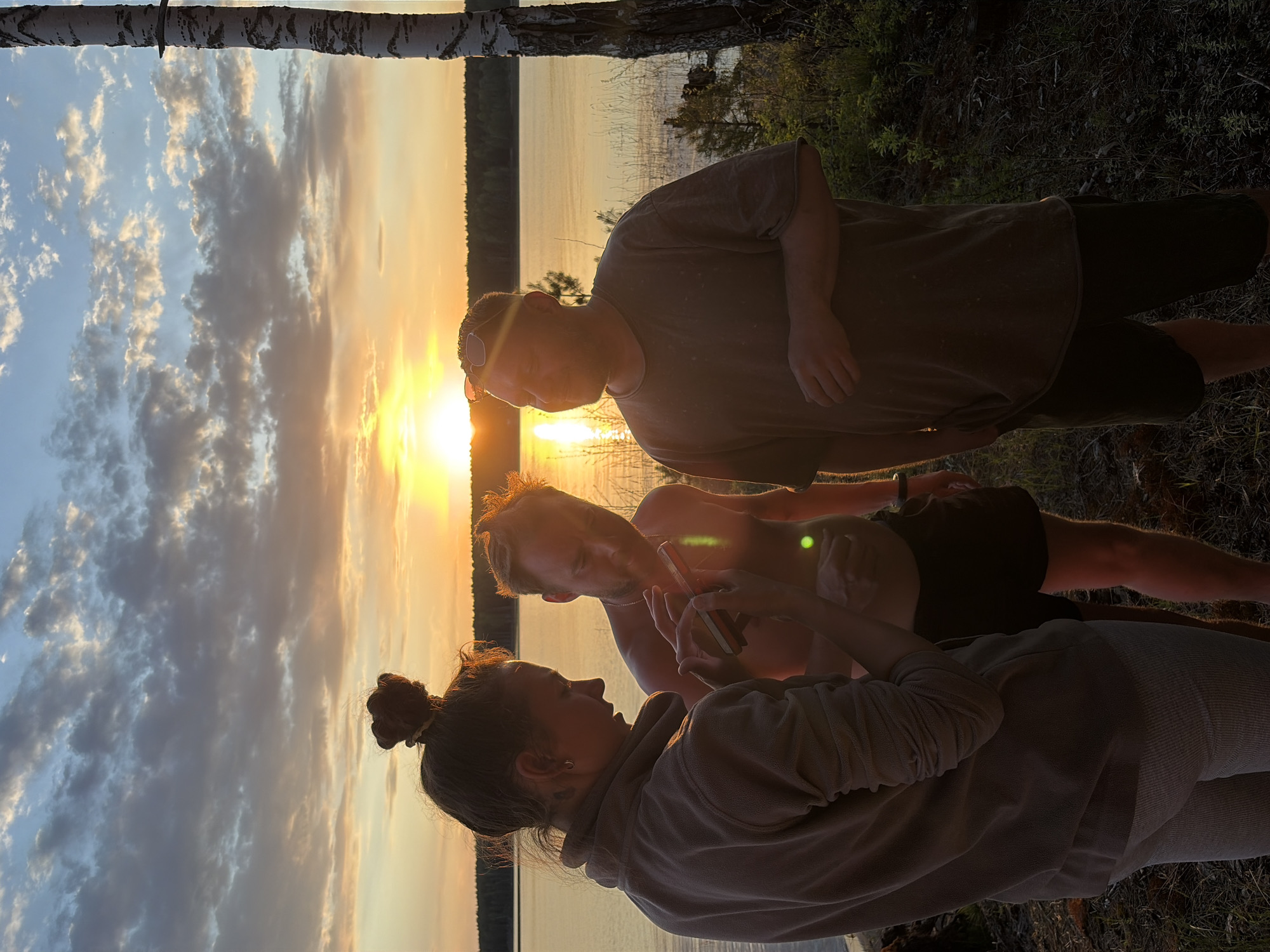 The image shows three individuals outdoors near a body of water. A woman with her hair in a bun is holding a mobile phone, appearing to show something to a shirtless man. A second man, wearing a t-shirt, is standing nearby. The background features a sunset reflected in the water. The scene appears to be taking place near or on a dock or pier. Trees are visible along the shoreline. The location is Zabegaevo, Russia.