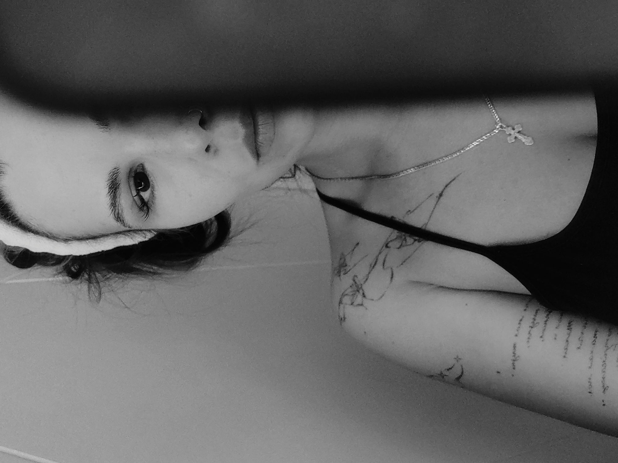 The image is a monochrome portrait of a person with pale skin and dark hair, possibly female, taken at an unusual angle. The person has a headband and is wearing a tank top. A silver necklace with a cross pendant is visible on their neck. The person has tattoos on their arms, including text. The location is unknown, and no evidence suggests this image was taken in the city of Chaikovsky, Russia.