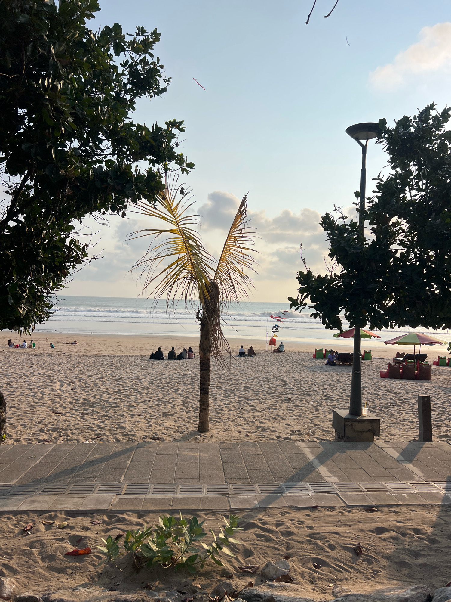 The image captures a serene beach scene in Kuta, Indonesia, during what appears to be late afternoon or early evening. The foreground features a textured sand path bordering the beach, with a few fallen leaves and small green plants adding natural detail. A gray concrete sidewalk runs alongside the sand, marked with square tiles and parallel drainage grates, casting long shadows from an unseen light source.
The beach itself is broad and sandy, with subtle footprints indicating recent activity. In the mid-ground, several groups of people are scattered across the sand. Some are seated, appearing to relax or socialize, while others are reclined, possibly sunbathing. A lone palm tree with sparse, windblown fronds stands prominently near the center, its trunk dried and weathered.
Further back, the ocean presents gentle waves rolling towards the shore. A few colorful kites are visible in the sky, adding a playful element to the scene. Along the edge of the beach, near the water, are some beach umbrellas and seating arrangements, suggesting a commercial area for beachgoers. Two tall, dark lampposts with modern, circular light fixtures stand on the sidewalk, one with a glass of yellow liquid resting on its base. The sky above is a soft blue, with scattered clouds and a bright, diffused light suggesting the sun is low on the horizon. The overall impression is one of tranquility and leisure on a popular Indonesian beach.