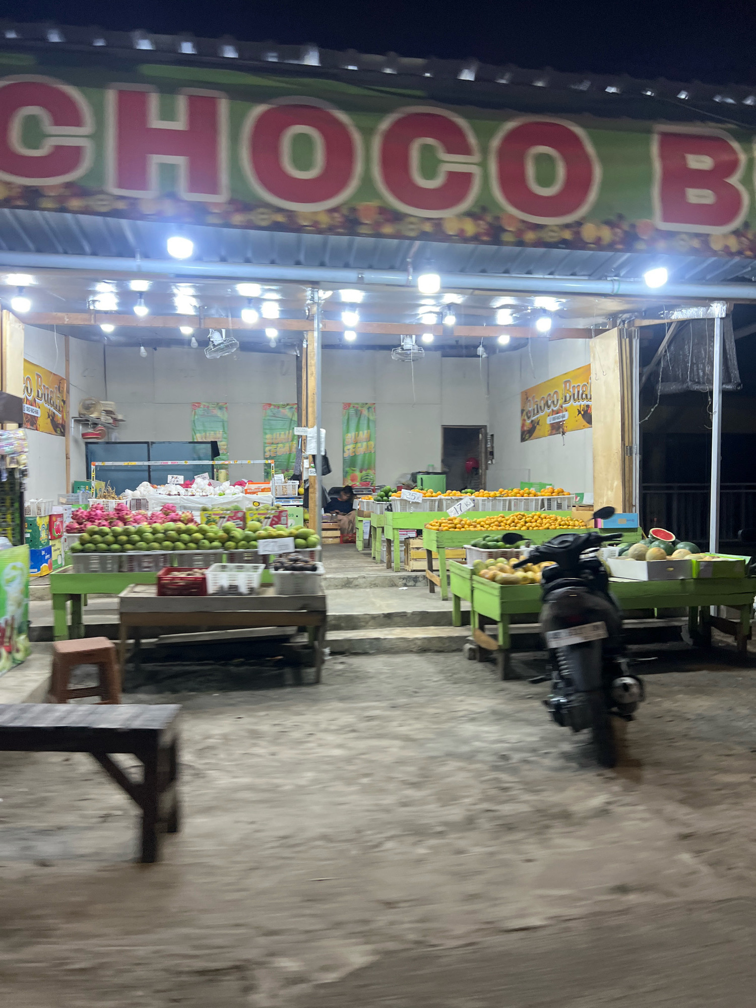 This image depicts a fruit stall located in the city of Payaman, Indonesia, at night. The stall features a prominent green banner with large red lettering that reads "CHOCO B." displayed above the entrance. The stall is illuminated by numerous overhead lights, casting a bright glow on the merchandise.
Various fruits are neatly arranged on green wooden tables, including stacks of dragon fruit, mangoes, oranges, watermelons, and other produce. Baskets and crates are used to display the fruits, and some have price tags attached, such as "Rp 25k" and "Rp 17k". In the foreground, a dark-colored motorcycle is parked on the uneven ground in front of the stall. A person is seated at a table to the right of the stall, facing away from the camera, and appears to be a vendor or customer. The overall environment suggests a busy roadside market.