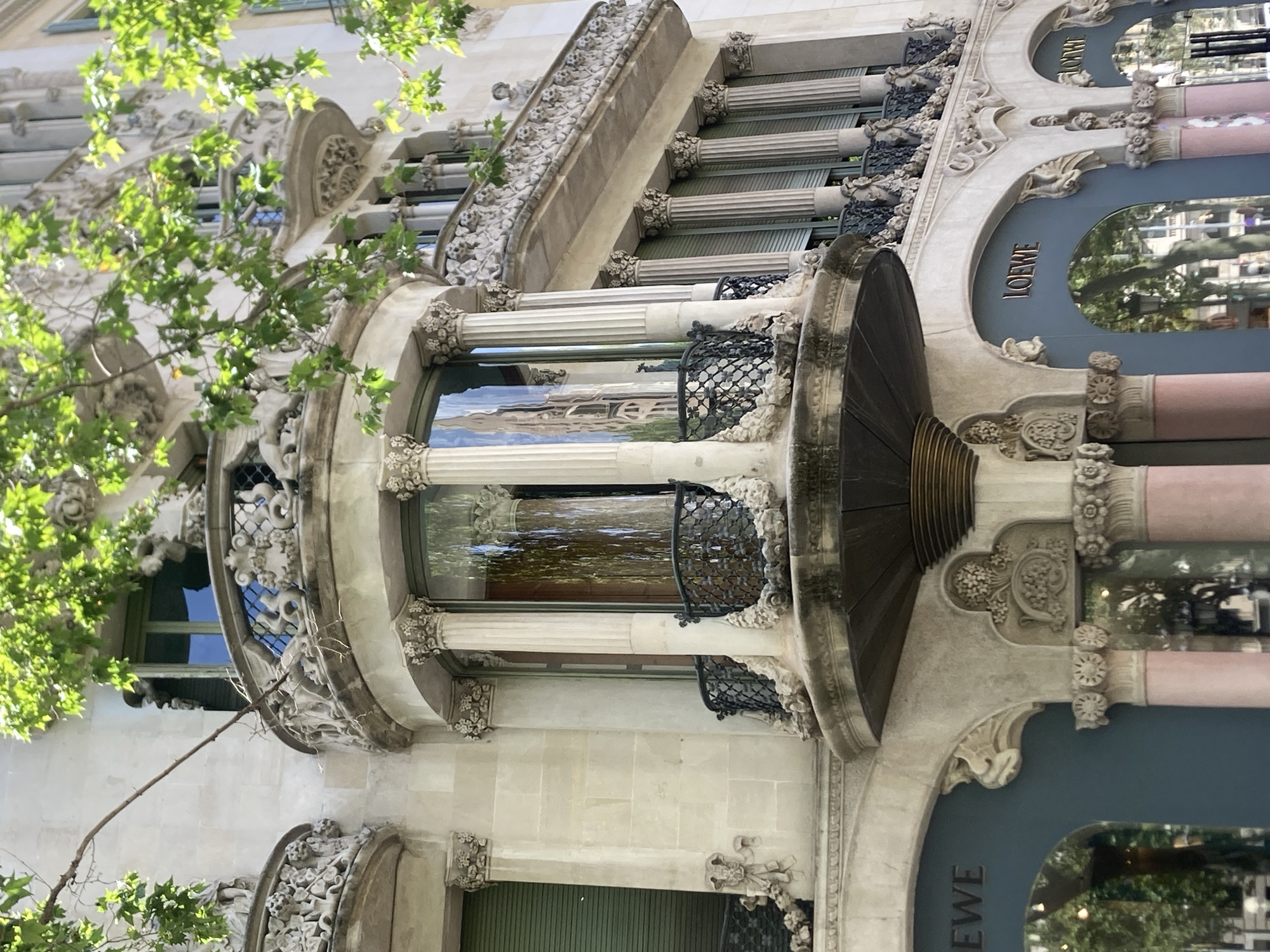The image shows the exterior of the Loewe store on Passeig de Gràcia in Barcelona, Spain. The building features ornate Art Nouveau architecture, with decorative balconies, columns, and stonework. Green leaves from a tree partially obscure the upper sections of the building. The Loewe logo is visible above the storefront windows, which reflect the surrounding streetscape and trees. The storefront is a deep teal color, contrasting with the lighter stone of the building's facade. The style of the architecture suggests it is a historic building.