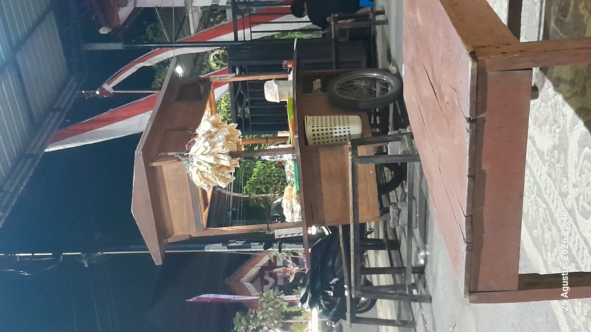 This is a nighttime, eye-level, medium shot of a street food stall in Madiun, Indonesia. The stall is made of wood with a tiered top and is supported by a cart with two large wheels. Hanging from the stall are numerous packets of food, likely snacks. Several bottles of drinks and a white waste bin are visible on the counter. In the background, a motorcycle is parked next to a table and benches. To the right, a person is seated at a table, facing away from the camera. Indonesian flags, red and white, are visible in the background. The ground appears to be paved with rough stones. The timestamp "25 Agustus 2026 22.48" is present in the lower left corner of the image.