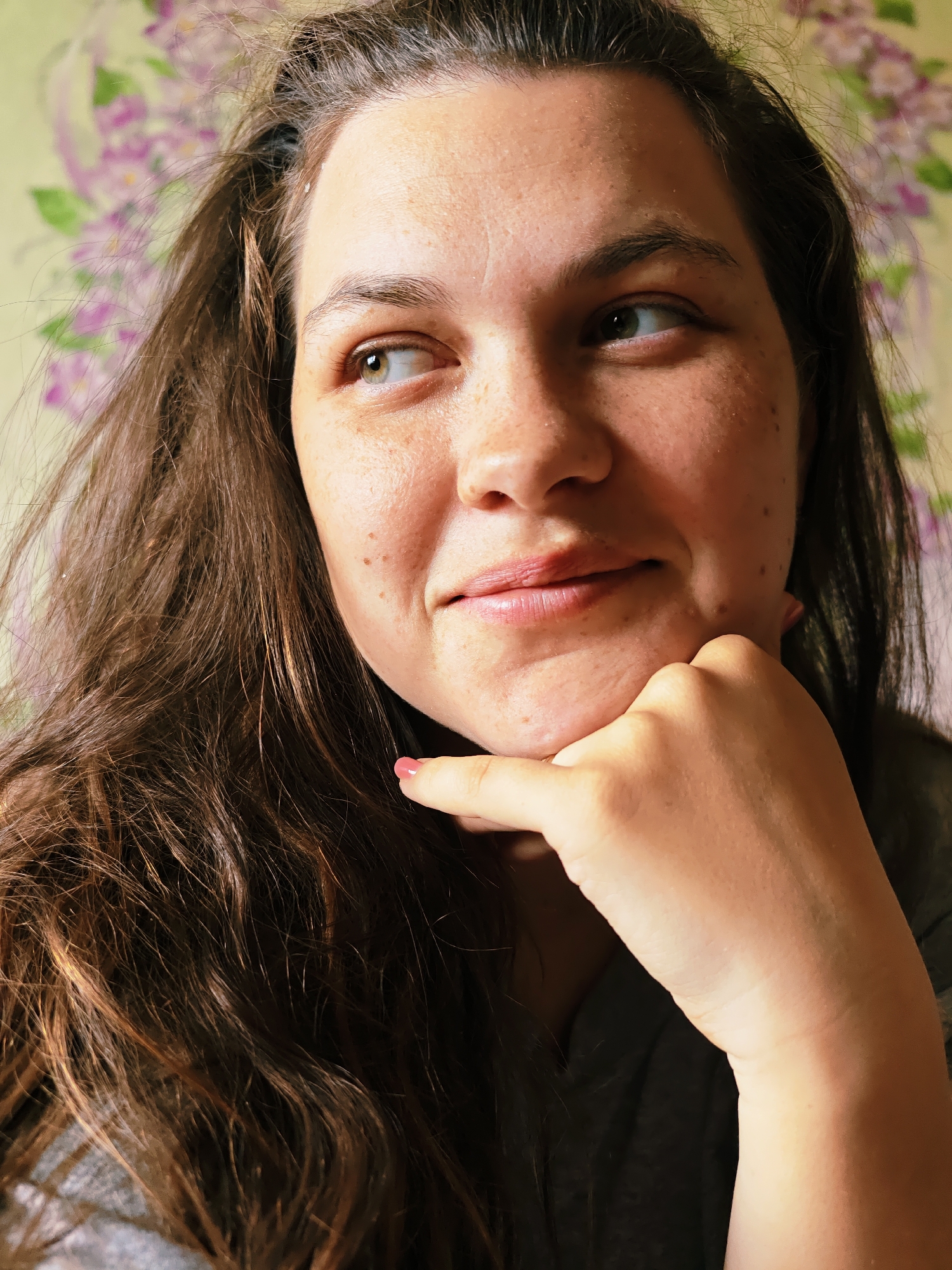 A close-up portrait shows a woman with long, dark brown hair and fair skin, adorned with numerous freckles. She has light-colored eyes, appearing green or hazel, and is looking slightly to her right with a subtle, gentle smile, suggesting a thoughtful or serene mood. Her right hand, featuring a pink-painted fingernail on the index finger, is resting under her chin. She is wearing a grey top or shirt. The background consists of a light green wall with a floral pattern depicting purple, lilac-like flowers, possibly wallpaper. The lighting indicates it is daytime. There is no visible text in the image. The provided location context is Уральское, Russia.