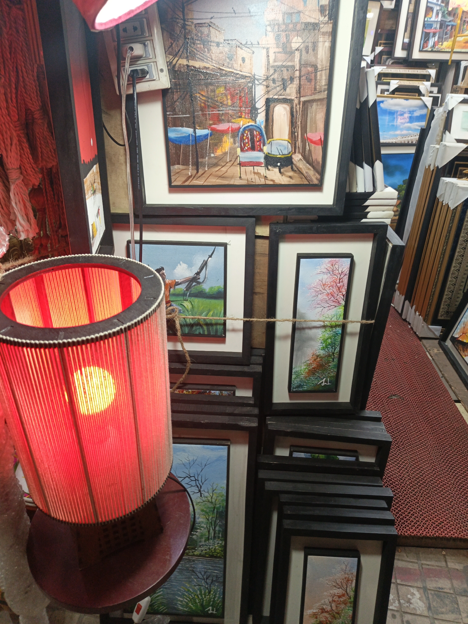 The image presents a vibrant, cluttered scene within an art stall or shop, likely in Dhaka, Bangladesh. A prominent, glowing red cylindrical lamp with vertical slats dominates the left foreground, its bright, warm light casting reflections and illuminating the immediate area. Its dark red base is visible below.
Behind the lamp and extending to the right, numerous framed paintings are stacked and displayed. The largest, at the top center, depicts a detailed street scene or alleyway with traditional buildings, overhead wires, and an empty, colorful rickshaw-like vehicle with blue and red coverings. Below it, another painting shows a figure, possibly a man, standing in a green field holding a long object, possibly a stick or rifle. To the right, a tall, narrow painting features a blossoming tree with pink and red leaves against a light blue sky, transitioning to green foliage at the bottom, bearing a signature "Jd." More framed artworks, including landscapes and scenes with clouds, are visible stacked behind and around these main pieces.
On the far left, a mass of red, textured material, resembling bundles of yarn or rope, hangs vertically. An electrical outlet with two cords plugged in is visible at the top left, with text "AWM 20276 80°C 30V VW-1 CABLE ARTWOOD" printed on one of the wires. The floor is covered with a distinctive patterned red rubber mat. The setting suggests an active commercial space, possibly a market stall, operating under artificial light.