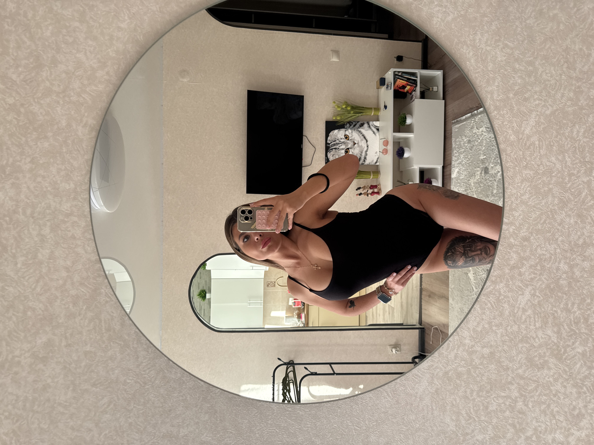 A woman is taking a mirror selfie in an apartment. She is wearing a black bodysuit and has tattoos on her thigh and arm. She is standing in front of a mirror that reflects the room behind her. The room appears to be a living room with a TV, a shelf with books and decorations, and a rug on the floor. The woman is holding her phone in her right hand and posing with her left hand on her hip. The image also shows a glimpse of a kitchen area through an arched doorway behind the woman. The apartment appears to be located in Sarapul, Russia, based on the information provided.