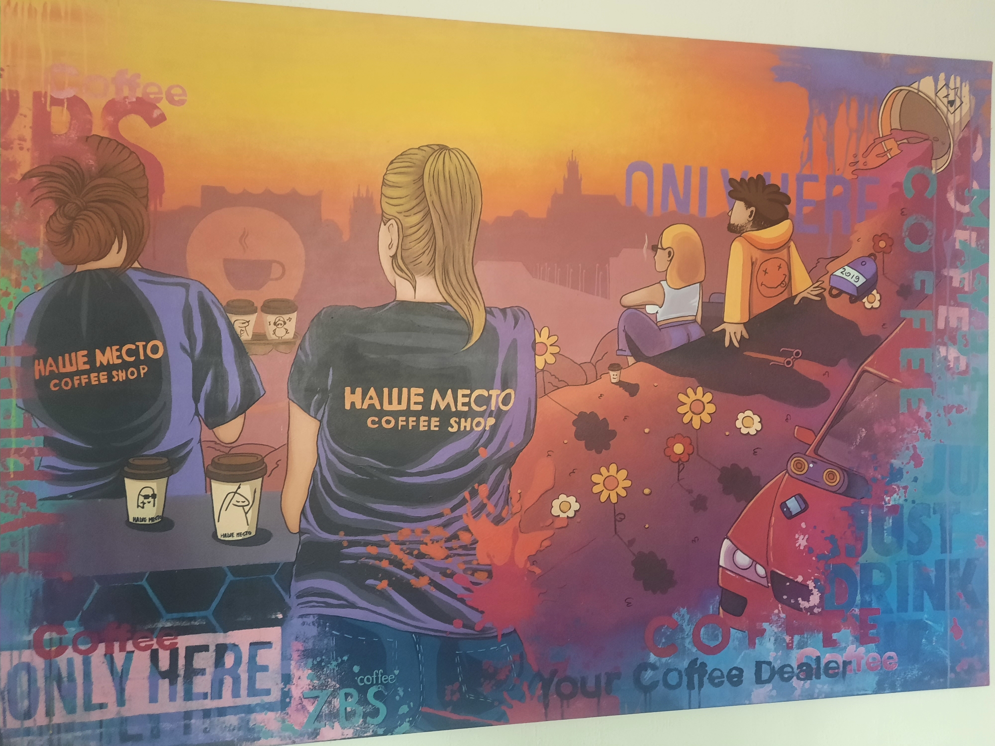 The image depicts a mural, likely painted on a wall in Hrodna, Belarus, with a vibrant and abstract style. The scene portrays several stylized figures against a sunset-like backdrop of an urban skyline.
In the foreground, two individuals are shown from behind, wearing t-shirts that read "НАШЕ МЕСТО COFFEE SHOP" (Our Place Coffee Shop). One has their hair styled in a high bun, and the other has a blonde ponytail. They are standing in front of two coffee cups, each adorned with a drawing of a stick figure.
Further to the right, two more figures are seated. One is a woman with blonde hair wearing glasses and smoking, and the other is a man with dark curly hair wearing an orange hoodie with a smiley face. A tipped-over coffee cup is pouring colorful liquid, resembling a paint spill, onto the scene.
The background shows silhouettes of buildings, suggesting a city. The mural is characterized by splatters of paint and graffiti-like text. Below the main figures, there are inscriptions that read "COFFEE ONLY HERE" and "Your Coffee Dealer". A red car is also depicted, partially integrated into the design. The overall color palette is warm, dominated by oranges, yellows, and purples, with splashes of red and blue. The mural appears to be an artistic representation promoting a coffee shop, possibly named "Our Place".