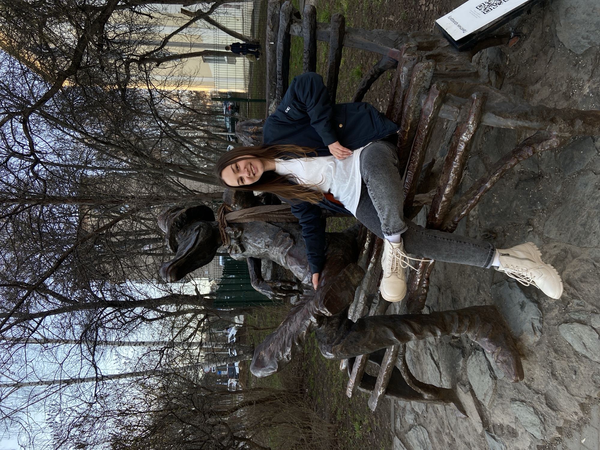 A young woman with long brown hair is smiling at the camera while sitting on a large, dark metal sculpture in an outdoor setting. She wears a white t-shirt, a dark blue jacket, grey jeans, and light-colored lace-up boots. Her expression suggests a cheerful and relaxed mood.
The sculpture is elaborate and abstract, appearing to be made of a dark, textured metal, possibly bronze or cast iron. It features intertwining forms that could represent figures or stylized organic shapes, and it functions as a unique bench or seating area.
To the right of the woman and sculpture, a white sign with a QR code is visible, resting on a rocky base. The Cyrillic text on the sign clearly reads "Памятник крокодилу," which translates to "Monument to the Crocodile."
The background is dominated by numerous bare trees with intricate branches, suggesting late autumn, winter, or early spring. Through the branches, patches of a pale blue sky are visible, indicating it is daytime, possibly late afternoon given a subtle warm tint in the upper sky. In the deeper background, elements of an urban environment can be discerned, including distant buildings, a fence, two faint figures, and a vehicle. The ground around the sculpture is a mix of earth and stones.