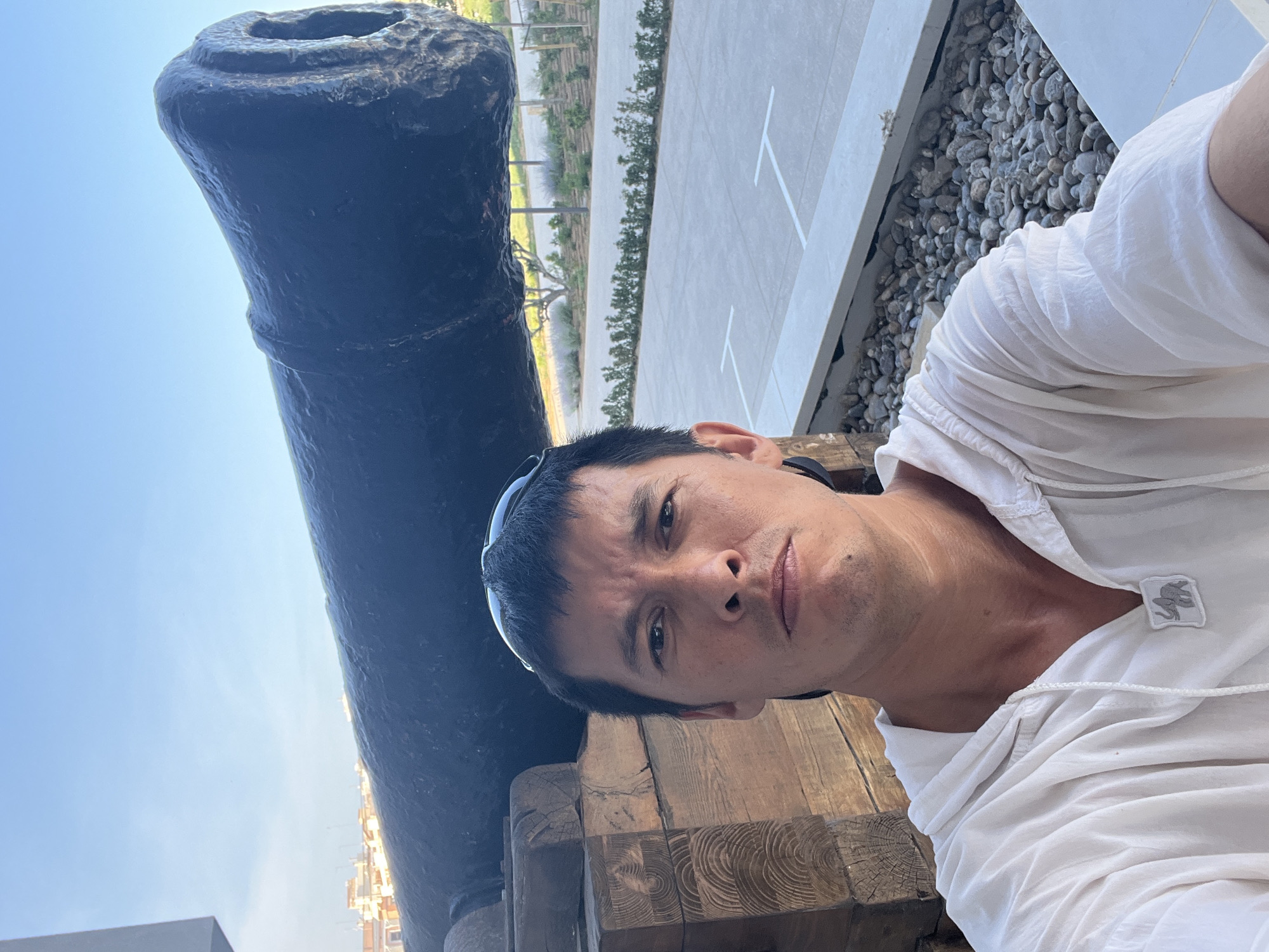 A man is taking a selfie outdoors, standing in front of a large, black cannon. He is wearing a white hooded shirt and sunglasses pushed up on his head. The cannon appears to be on display, supported by wooden beams. Behind the cannon, there is a paved area with some landscaping and buildings visible in the distance. The sky is clear and blue. This scene is likely located at the historical fort of Tamarit, located in the municipality of Altafulla, near the city of Tarragona, Spain.