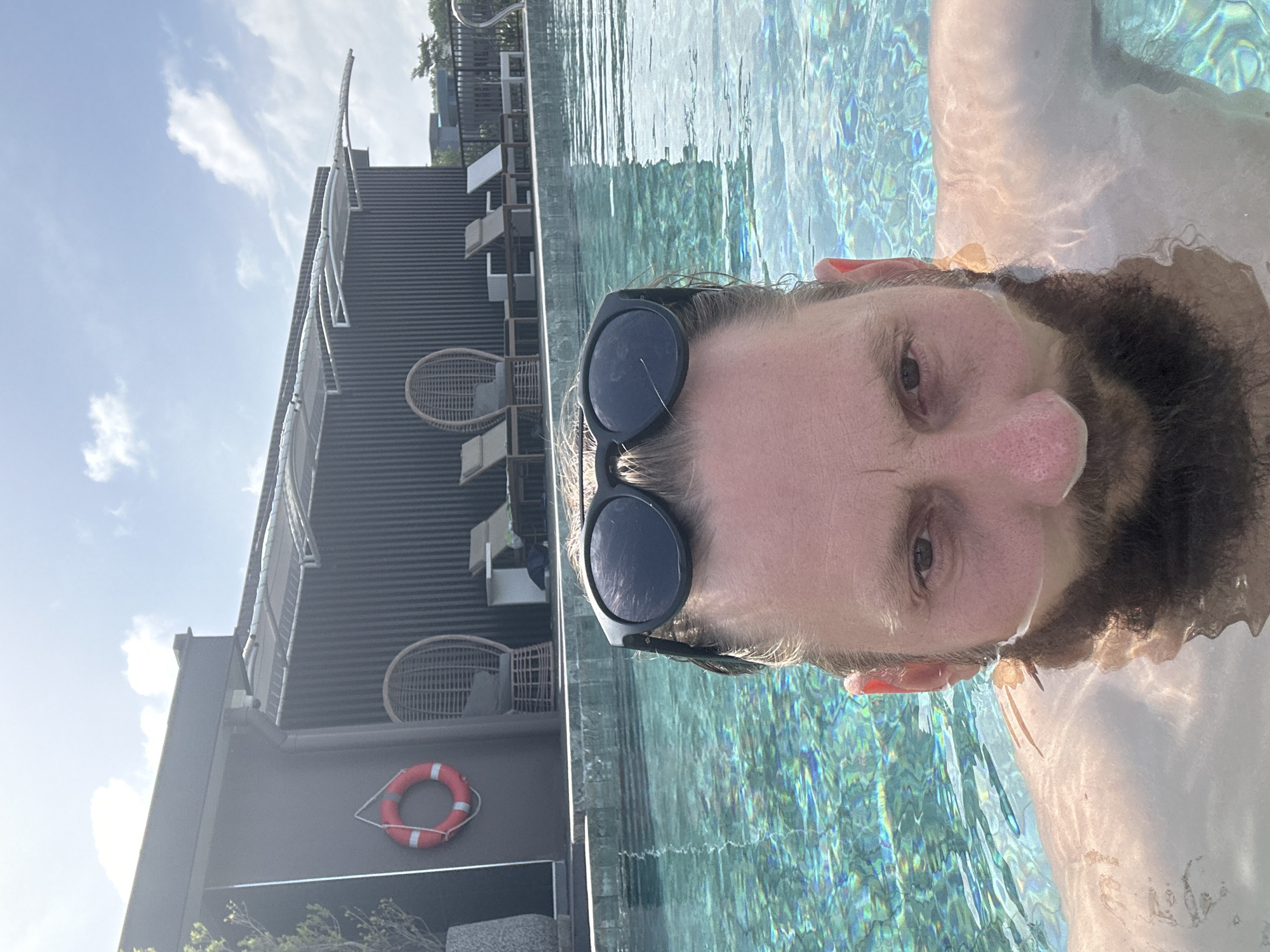 In Rawai, Thailand, a person is in a swimming pool, taking a selfie. The person is wearing black sunglasses and has a beard and short brown hair. Part of their body is submerged in the clear blue water. In the background, there is a building with a dark corrugated metal facade and a roof structure with white fabric canopies. A red life preserver is hanging on the side of the building. There are lounge chairs and small tables arranged near the pool. The sky is blue with scattered white clouds, indicating a sunny day.