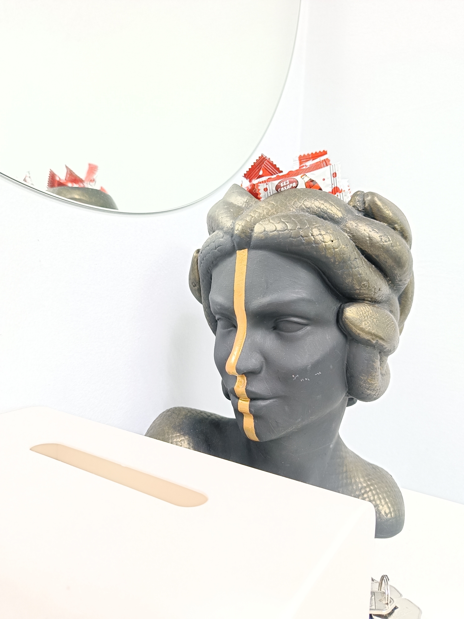 An indoor, brightly lit scene features a decorative bust of Medusa, rendered in dark gray with gold accents. The bust, depicted from the chest up, has gold-colored snakes for hair and a distinctive vertical gold stripe painted down the center of its face, from the forehead to below the nose. Numerous red and white candy wrappers are arranged on top of the Medusa bust, nestled among the snake-like hair, suggesting it is being used as a candy holder. One wrapper clearly displays the Russian text "БЕЗ САХАРА," meaning "SUGAR-FREE."
In the upper left background, a round mirror reflects a portion of the candy wrappers and the top of the bust. The wall behind is plain white, contributing to a clean and minimalist aesthetic. In the foreground, a light-colored rectangular object, possibly a dispenser or a counter section, partially obscures the lower part of the Medusa bust. This object has a horizontal, rounded slot on its top surface. A set of keys on a metal ring is partially visible in the bottom right corner, resting on a white surface. The scene is static, devoid of human presence or activity, and is likely set in a modern office, clinic, or reception area in Chaikovsky, Russia. The bright, even lighting suggests either strong artificial light or a well-lit interior during the daytime.
