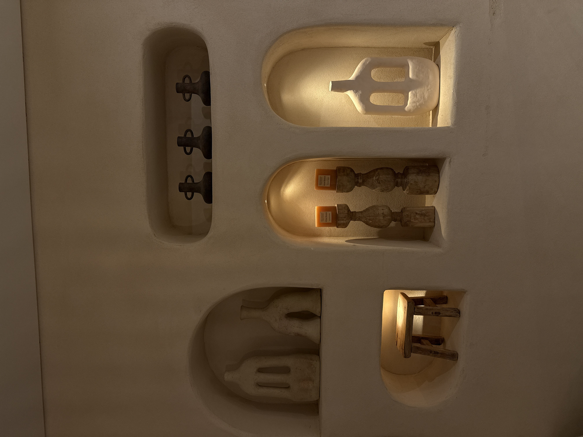 This image shows a close-up of a textured, off-white wall with several recessed alcoves, possibly in a home or a retail space in the Tamar Regional Council, Israel. The alcoves are illuminated by warm, ambient lighting, highlighting the decorative objects within.
In the upper right alcove, three dark, minimalist vase-like objects with loop handles are displayed. Below this, in a central alcove, are two tall, carved wooden candle holders, each topped with an orange-colored candle. To the right of these candles, in another alcove, sits a large, abstract, off-white sculpture with geometric cutouts, resembling a stylized human figure.
To the left of the candle holders, in a larger alcove, are two off-white, abstract sculptures. The sculpture on the left is taller and more rectangular with two vertical slits, while the one on the right is shorter, wider, and has a more curved, organic shape with an archway.
In the lower left, a small, rustic wooden stool with four legs is placed inside a round alcove, also illuminated by a warm light. The overall aesthetic is minimalist and earthy, with a focus on texture and form. No people are visible in the image.