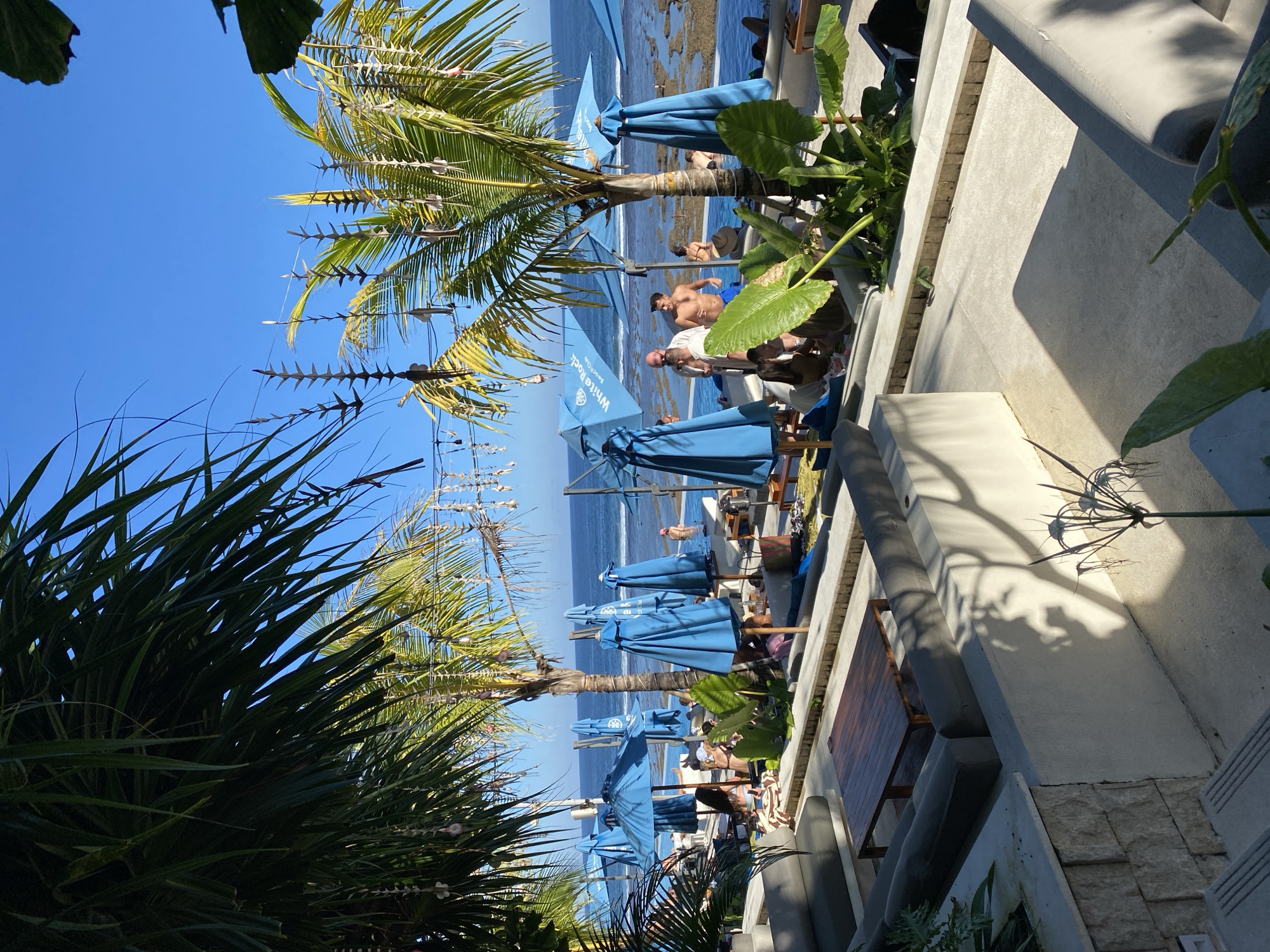 This media file depicts a beachside scene with several people relaxing and enjoying the outdoors. The setting is a resort or restaurant with lounge chairs and blue umbrellas, suggesting a leisure activity. The ocean is visible in the background, with calm water and a rocky shoreline. Palm trees are prominent in the foreground, framing the scene and adding to the tropical ambiance.
Several individuals are visible in various states of relaxation, some seated and others standing. One person appears to be a server, tending to guests or preparing the area. The overall mood seems peaceful and relaxed, indicative of a vacation or a leisurely day at the beach.
The time of day appears to be daytime, with bright sunlight casting shadows, suggesting it's likely midday or early afternoon. The weather seems pleasant, with clear blue skies and calm seas. There are several blue umbrellas open, providing shade.
Visible text on one of the umbrellas reads "White Rock Beach Club". This, combined with the visual cues of palm trees, ocean, and beach, strongly suggests a tropical beach location, consistent with Kuta Selatan, Indonesia. The architecture of the resort, with its concrete structures and wooden accents, is also characteristic of many beach resorts in the region.