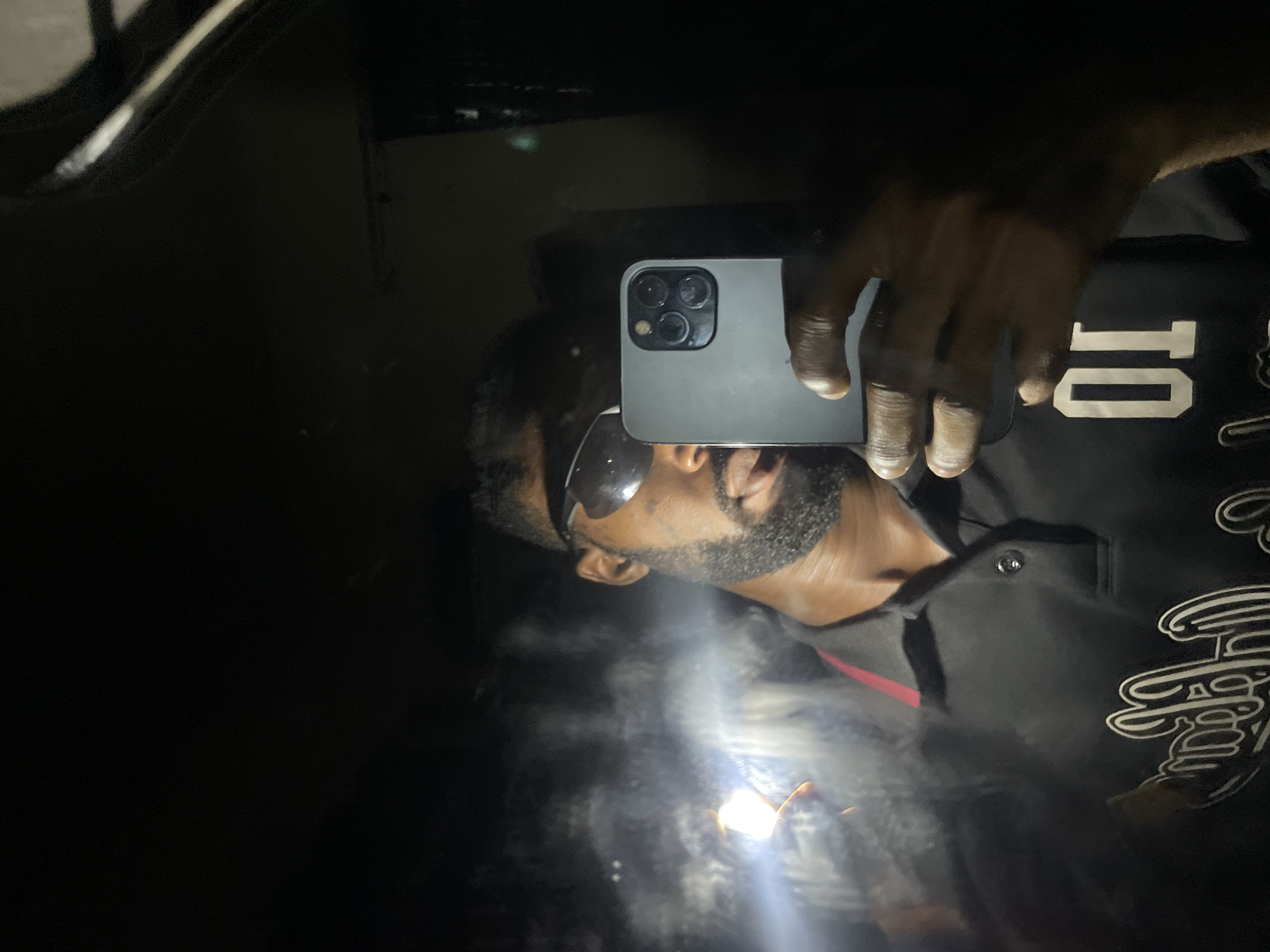 A male individual with dark skin, a beard, and short hair is depicted in a mirror selfie taken in Lokoja, Nigeria. He wears dark, reflective sunglasses and a black polo shirt featuring a red collar stripe and white stylized lettering, including the prominent number '01' on the chest. The subject holds a gray iPhone, identifiable by its triple-lens rear camera system, in his right hand, positioned to capture the reflection. His left hand grasps a small, intense light source that emits a bright beam, illuminating visible vapor or particulate matter in the air to his left. The surrounding environment is dark and characterized by reflective surfaces, indicative of a dimly lit interior with minimal ambient light.