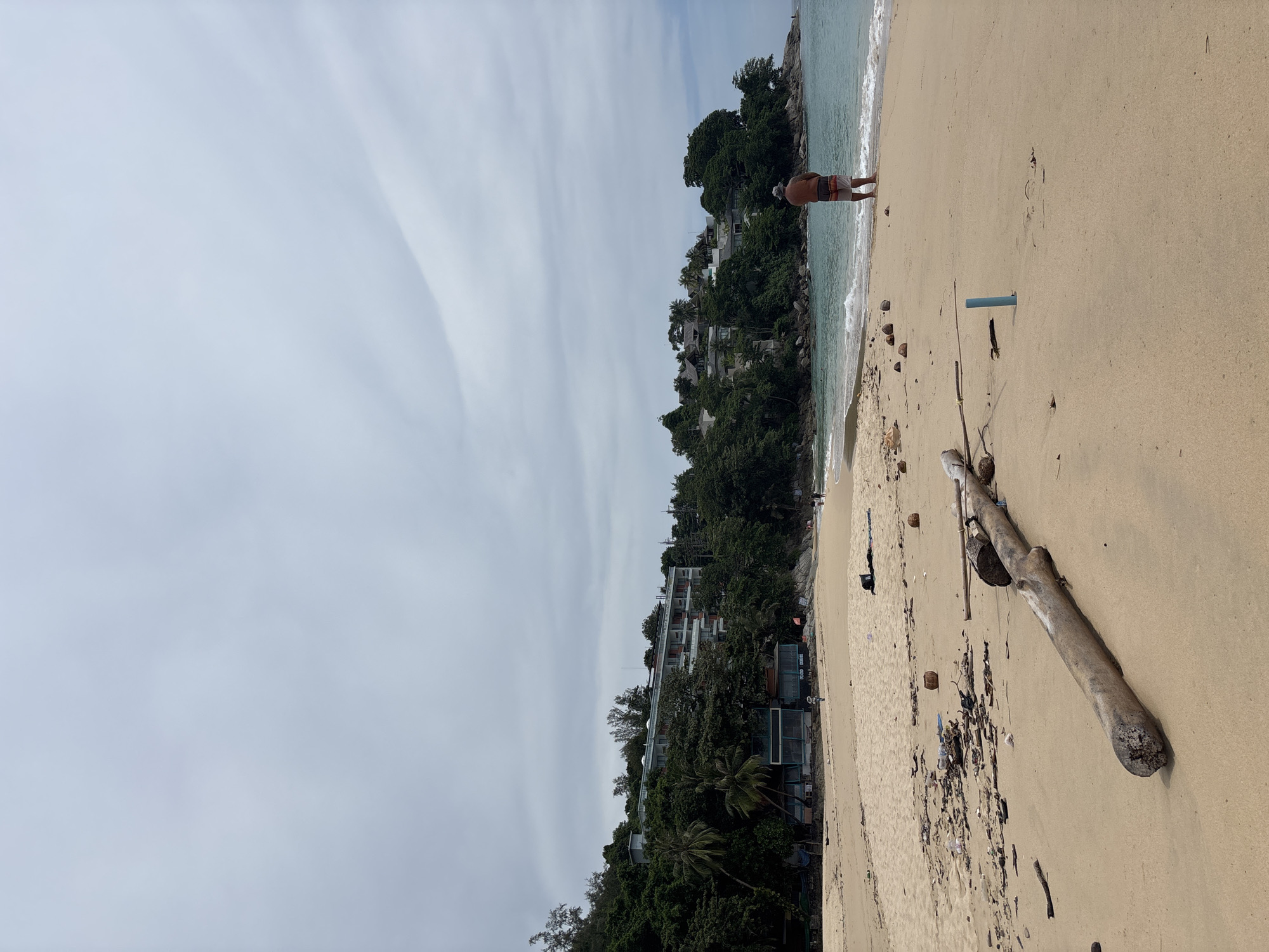 The image shows a beach in Choeng Thale, Thailand. The scene is oriented with the shoreline running vertically on the right side of the frame. A man stands at the edge of the water, facing away from the camera. The water is light blue and the sky is overcast. The beach is sandy and contains a long, weathered log. There is also trash, including plastic bottles, on the sand. Further down the beach, there are buildings and lush tropical vegetation on a hill overlooking the beach.