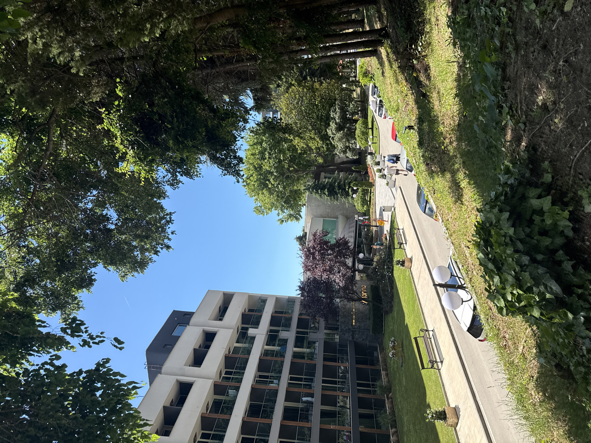The image depicts a view of the Marina residential complex in Varna, Bulgaria, on a clear, sunny day. In the foreground, a grassy embankment slopes downwards, covered in lush green vegetation and shadowed by tall trees lining the right side of the frame. A paved pathway runs alongside a road, bordered by a manicured lawn. Several cars, including a white hatchback and a red sedan, are parked on the road. Two white streetlights with spherical globes stand by the side of the road.
Dominating the left side of the image is a modern, multi-story building with a white facade, featuring numerous balconies with glass railings. A dark grey section is visible at the top of the building. Adjacent to this building, a sign with the word "marina" in golden lettering is affixed to a stone facade. In front of the building, a small garden area features benches and decorative planters.
Further back, more trees and greenery are visible, along with another building with a curved facade and green-tinted windows. Several people are walking along the pathway, some dressed in casual summer attire. The sky is a vibrant, cloudless blue, with a faint contrail from an aircraft visible in the upper left quadrant. The scene conveys a sense of a peaceful, well-maintained urban environment.
