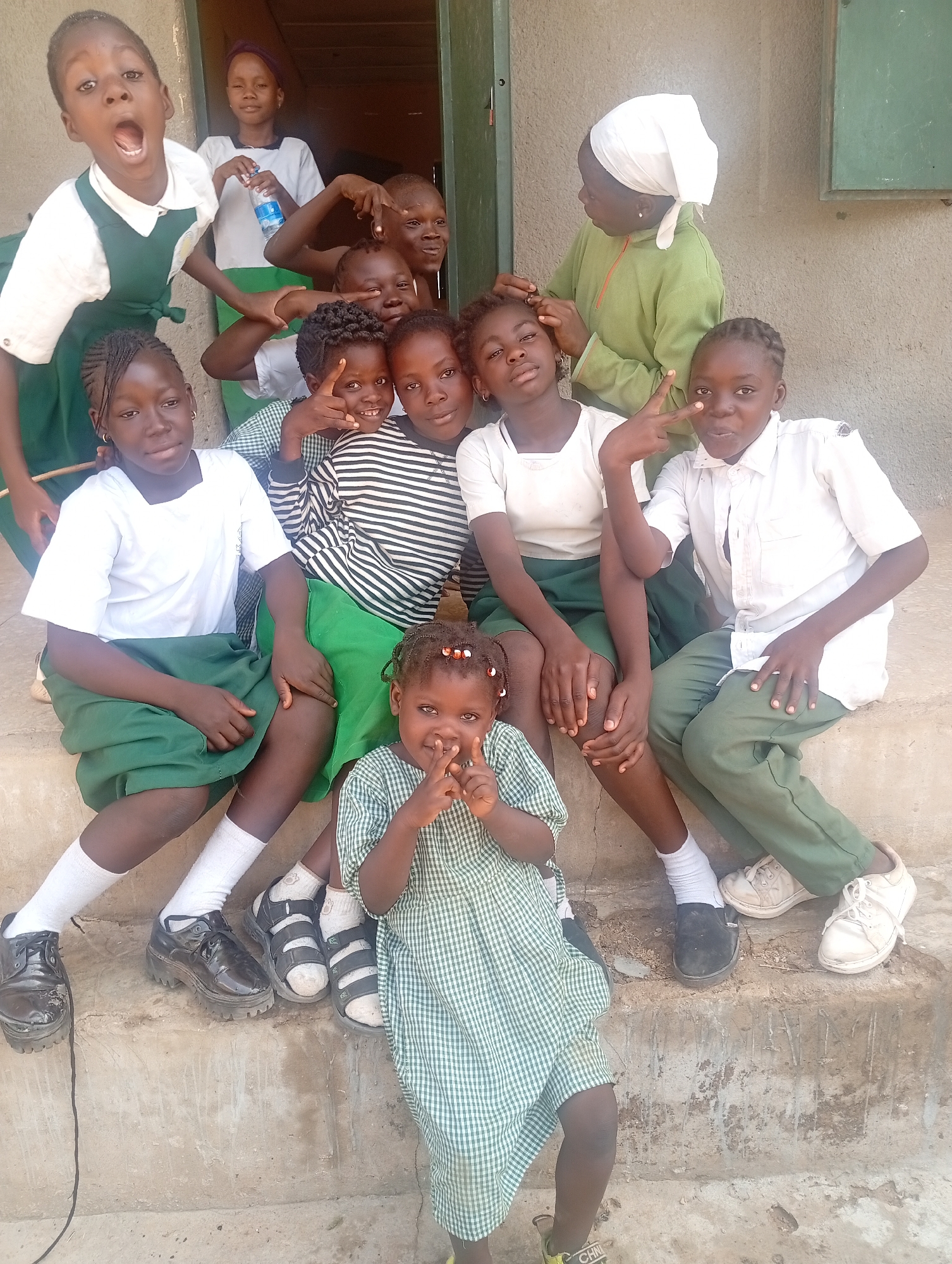 A group of children are gathered on the steps of a building in Narayi, Nigeria. Most of the children are wearing school uniforms with white shirts and green skirts or pants. One child is wearing a green and white checked dress and another is wearing a striped shirt. A woman in a green garment and a white headscarf is tending to the hair of one of the girls. Several of the children are making hand gestures, including peace signs and finger guns. The building is a simple structure with a door and a window. The steps are made of concrete.