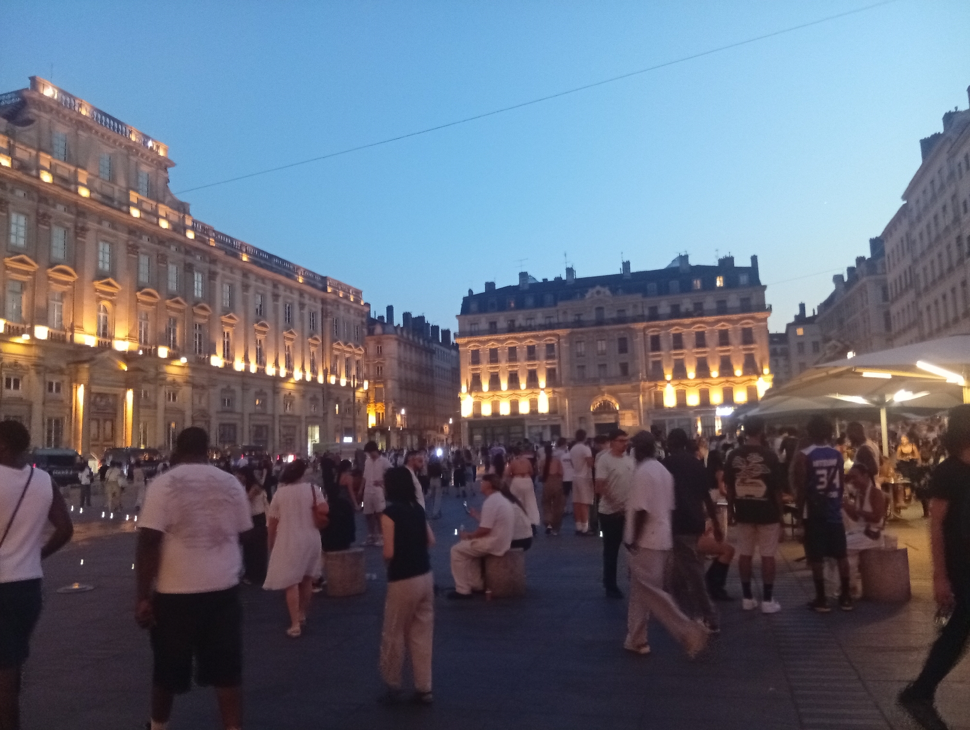 The image captures an evening scene in Lyon, France, likely at Place Bellecour, a large public square. The sky is a clear twilight blue, with the warm glow of artificial lights illuminating the grand, ornate architecture of the surrounding buildings. Several buildings are visible, characterized by their neoclassical style, with multiple stories, large windows, and decorative facades. The building on the left is particularly prominent, featuring a grand entrance and illuminated upper floors.
In the foreground and midground, a diverse crowd of people are gathered, suggesting a lively atmosphere. Many individuals are casually dressed, and their activities appear varied: some are walking, others are conversing, and a few are seated on stone blocks, possibly resting or socializing. The presence of tents or canopies in the background suggests a market or an outdoor event. The ground is paved, typical of a public square. The overall impression is one of a bustling urban environment transitioning from day to night, with people enjoying the open space and the illuminated architecture.