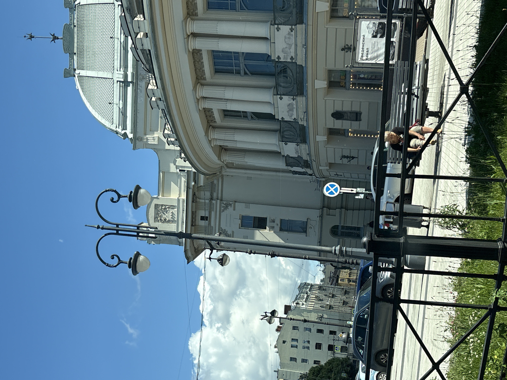 The image captures a street scene in Saint Petersburg, Russia, likely during daytime. In the foreground, a black metal fence with crisscrossing bars separates the viewer from the rest of the scene, with green grass and small white flowers growing beneath it. A woman with blonde hair is seated on a park bench, facing away from the camera and towards a building. She is wearing a dark t-shirt and light-colored shorts. She appears to be looking down at her hands. Behind her, a large, ornate building with a domed roof and classical architectural elements, including columns and decorative detailing, dominates the right side of the frame. A street lamp with two globes stands tall to the left of the building. In the background, several other buildings of varying architectural styles are visible, along with a clear blue sky with scattered white clouds. A street sign with a blue circle and a white cross is visible on a pole. Several cars are parked or driving on the street. A billboard with Russian text and an image of a canal is also visible.