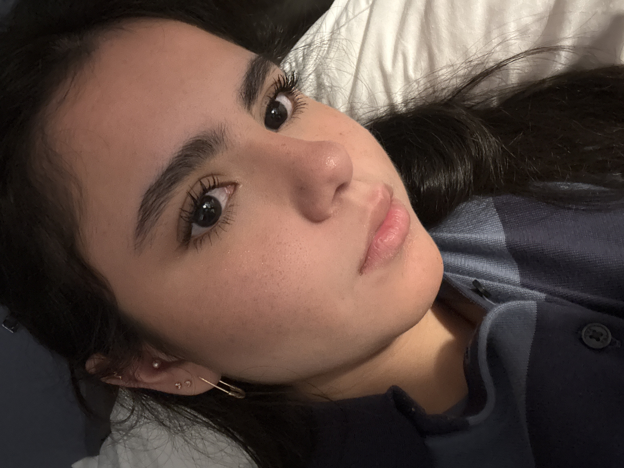 A close-up photograph depicts a woman lying down, possibly on a bed, with a white pillow visible behind her head. She is facing towards the camera with her head tilted slightly. Her dark hair is spread out around her. She has dark eyebrows, dark eyelashes, and clear skin. She is wearing earrings and a safety pin earring. She is wearing a blue plaid shirt. The image does not contain enough information to establish a precise location, particularly in Chicago.