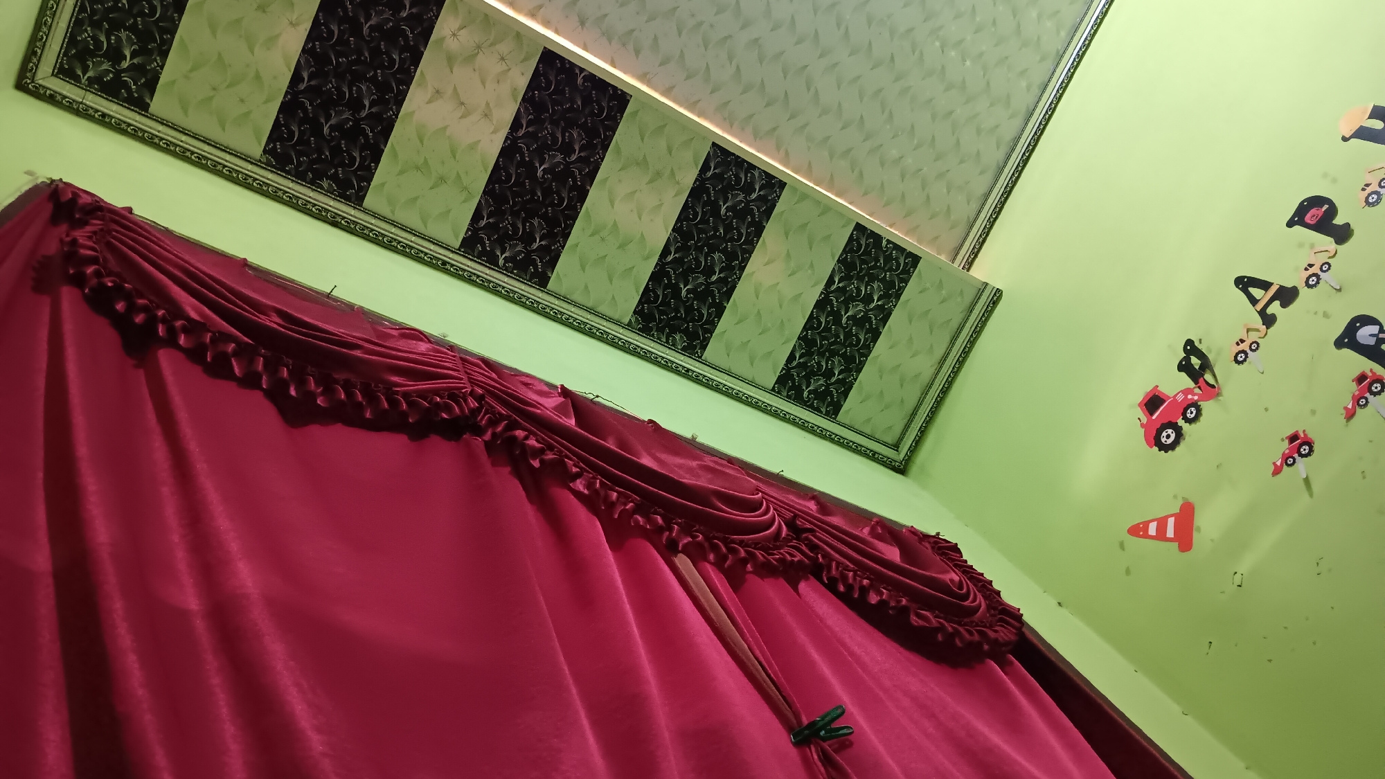 The image depicts a room with a light green wall and a ceiling with a patterned design. In the foreground, a rich crimson curtain with decorative ruffles drapes across the frame. The ceiling features alternating panels of light green and dark grey with intricate white patterns. On the light green wall, several colorful wooden cutouts of vehicles, including red trucks and a traffic cone, are affixed. The perspective is upward, suggesting the photo was taken from a low angle, looking towards the ceiling. The scene appears to be a child's room. The location is identified as Sonobekel, Indonesia.