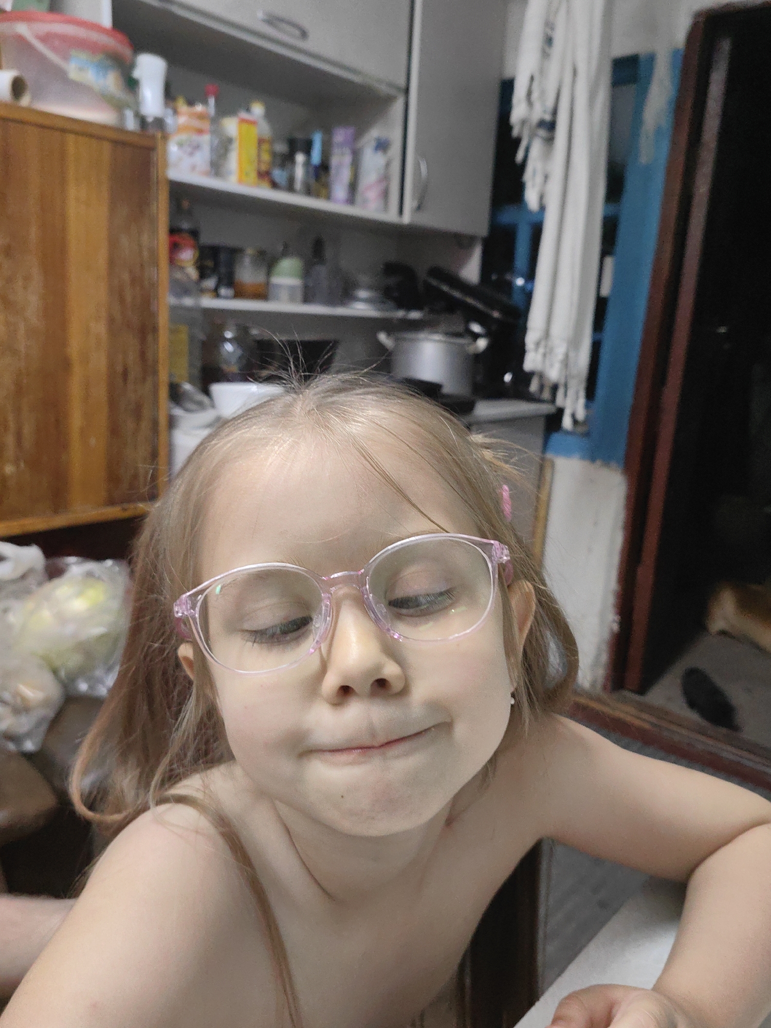 This media file is a close-up, eye-level photograph of a young girl wearing pink-rimmed glasses. Her eyes are closed, and she has a slight smile on her face. Her hair is light brown and appears to be tied back with a pink accessory. The girl is bare-chested, and her skin is lightly tanned.
The background is blurred, but it appears to be a domestic setting, possibly a kitchen. To the left, a wooden cabinet is visible, and shelves with various items, including containers and bottles, can be seen. In the mid-ground, a metallic pot is present, and to the right, a dark doorway or opening is visible, with a hint of a lighter-colored surface and a blurry, light-brown shape that could be an animal. A white curtain hangs in the background on the right side.
The lighting suggests it could be daytime, possibly indoors, as there are no strong indications of natural outdoor light. The overall mood conveyed by the girl's expression is one of contentment or playful silliness. There is no visible text in the image.