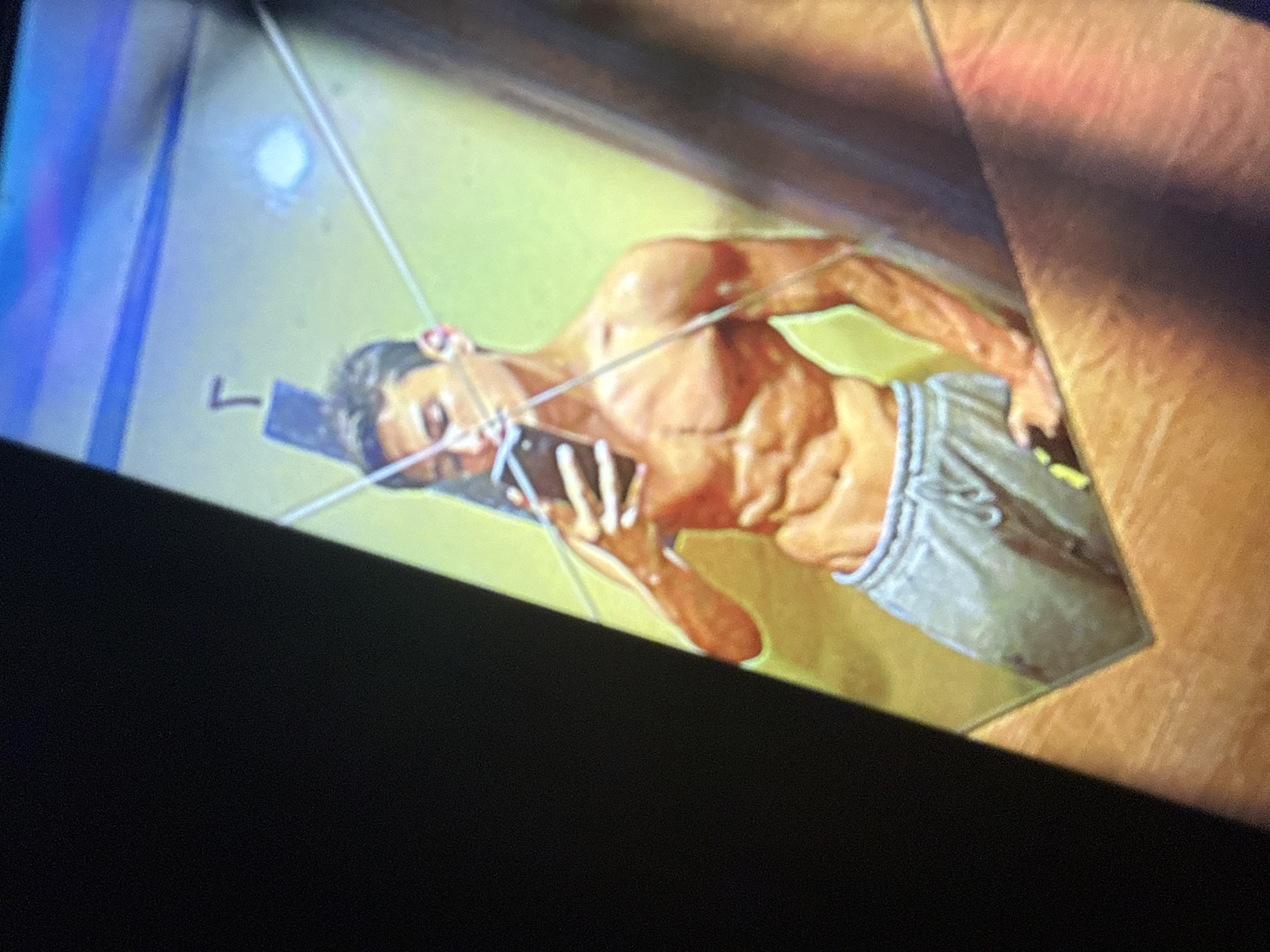The image shows a young man with well-defined abdominal muscles, wearing gray sweatpants, taking a mirror selfie. He is holding a smartphone in his right hand, angled to capture his reflection. The background appears to be a room with a lime green wall and a blue ceiling. There are some reflective surfaces, possibly a mirror and a window, with diagonal lines across the image, indicating possible damage or distortion. There is a subtle 'Γ' symbol visible on the wall behind the man. The overall lighting is warm, with highlights on his skin. There is no discernible information in the image that would allow for determining the location within the city of Sofia, Bulgaria.