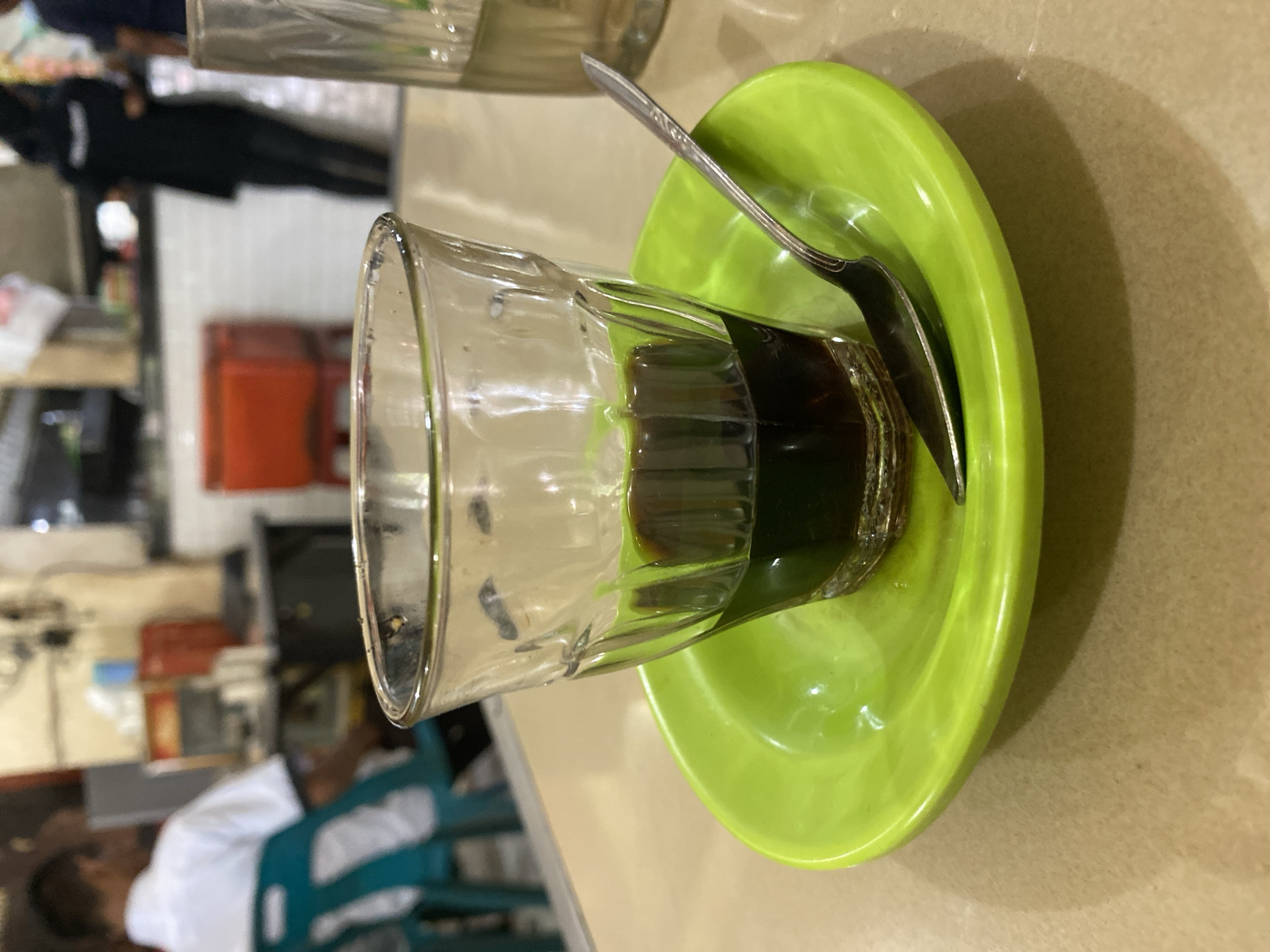 A close-up, eye-level shot focuses on a glass of dark liquid, likely coffee, resting on a bright green saucer. A small silver spoon lies on the saucer, its handle pointing towards the glass. The glass is about a third full, and the liquid has a glossy sheen. The saucer has a slight indentation in the center.
In the blurred background, a person wearing a white shirt is seated, facing away from the camera. To their left, a teal-colored chair is visible. Further in the background, other people are blurred, one wearing a black headscarf and dark clothing, and another figure to the far right. The environment appears to be an indoor establishment, possibly a cafe or restaurant, with a counter and shelving visible. The overall lighting is dim, with some bright spots reflecting off the glass and saucer. The image was captured in Lhokseumawe, Indonesia, suggesting a local eatery setting.