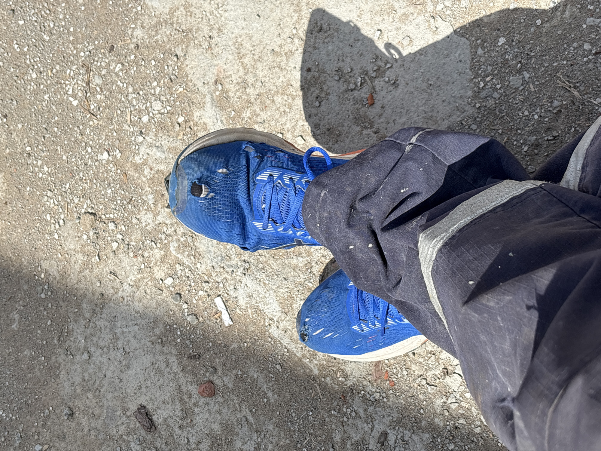The image presents a top-down view of a person's lower legs and feet, likely taken outdoors in Chaikovsky, Russia. The individual is wearing a pair of well-worn, bright blue athletic shoes with white soles and blue laces. Both shoes show significant wear, particularly with holes visible in the toe area of each shoe, suggesting heavy use or damage.
The person is also wearing dark blue work trousers or overalls, which appear durable and are splattered with light-colored specks, possibly paint, dust, or concrete. A reflective strip is visible on the outside of the right leg, characteristic of workwear. The trousers are somewhat creased and show signs of use.
The ground beneath the feet is a rough, textured surface, appearing to be concrete or asphalt mixed with small pebbles, gravel, and dirt. Scattered debris, including what looks like a cigarette butt, small pieces of brick or stone, and dry organic matter like twigs, is visible on the ground.
Strong, distinct shadows are cast by the person's legs and feet, indicating bright sunlight and suggesting it is daytime. The overall impression is one of a working environment or a casual outdoor setting where utility and wear are evident in the clothing and footwear. There is no visible text in the image. The activity appears to be simply standing or pausing.