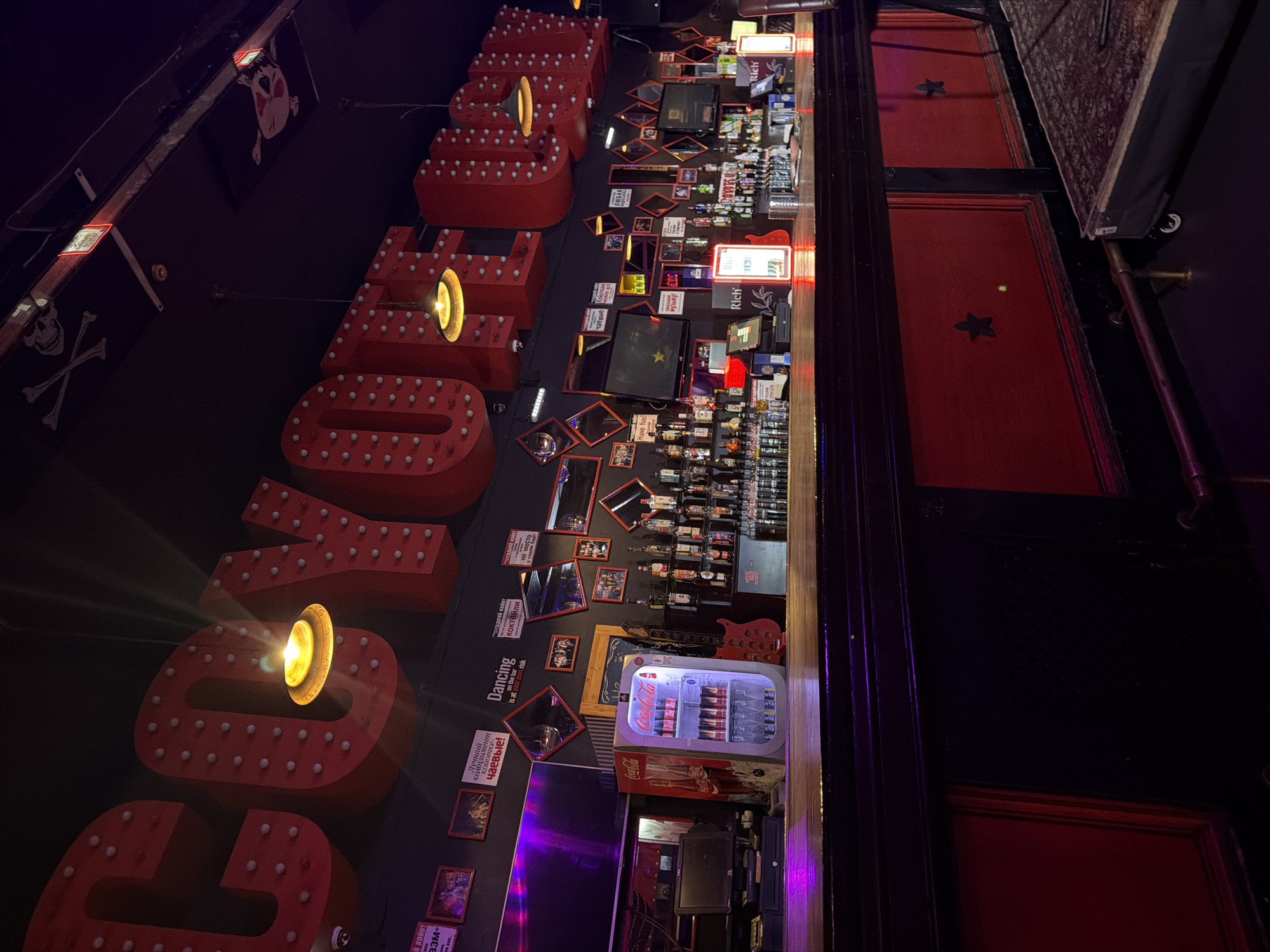 The image depicts the interior of a dimly lit bar or nightclub, identified as "Coyote Ugly" (Гадкий койот) in Kazan, Russia. The setting suggests an evening or nighttime environment with an energetic, edgy theme dominated by dark tones and red accents.
On the left, large, red marquee-style letters with embedded light bulbs spell out "Гадкий койот" (Coyote Ugly) against a dark wall. One letter features a prominent spotlight-like glow. Above these letters, pirate-themed banners with skull and crossbones designs are visible, enhancing the bar's distinctive decor.
The central wall area is adorned with various framed pictures, small mirrors, and informational signs, many in Russian. Key text includes "Dancing on the bar is at your own risk" (in English), and "Лучший комплимент койотке - чаевые!" (The best compliment for a coyote girl is tips!), which directly reference the bar's famed concept. Other Russian text seen includes "коктейли" (cocktails), "чаевые" (tips), "музыку!" (music!), and "ЛЮБАЯ" (any). A retro Coca-Cola mini-fridge filled with beverages and a red guitar cut-out are also featured on this wall.
To the right, a well-stocked bar counter displays numerous bottles of alcohol and illuminated "Richi" brand signs. Beyond the bar, a dark red paneled wall with black star decorations completes the thematic styling. Although no people are visible, the setup, signage, and extensive liquor display strongly imply a vibrant nightlife activity, characteristic of a bar known for dancing and entertainment.