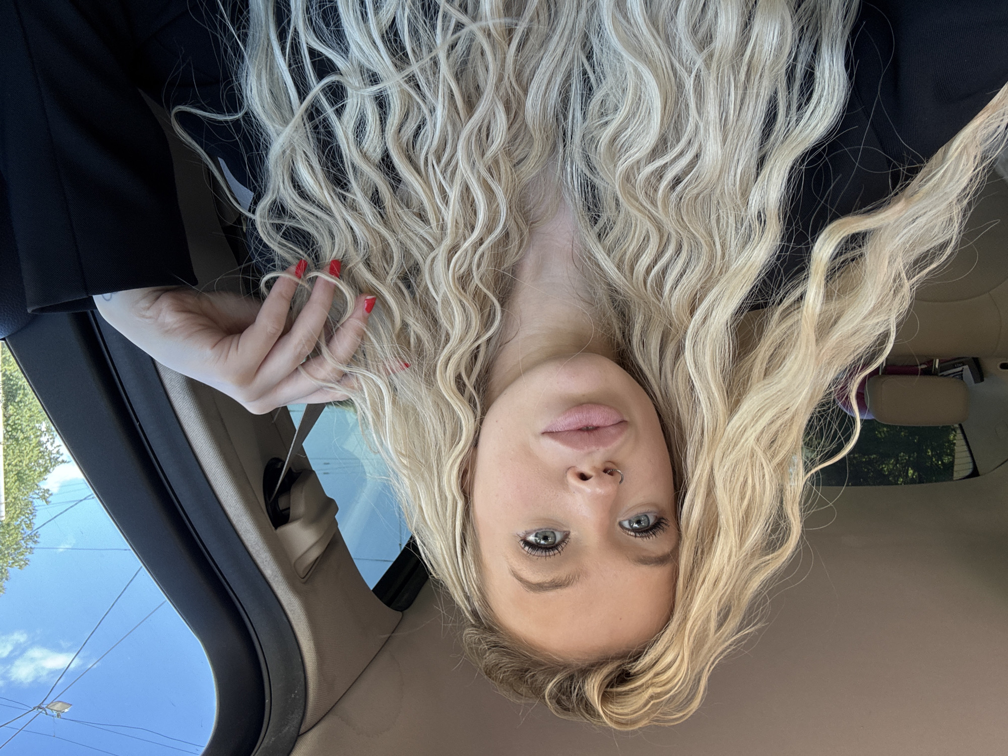A blonde woman with long, curly hair is sitting in the passenger seat of a car. She is wearing a black jacket. Her right hand is resting on her hair. The interior of the car is visible, as is the sky and trees through the window. The location is not Saint Petersburg, Russia.