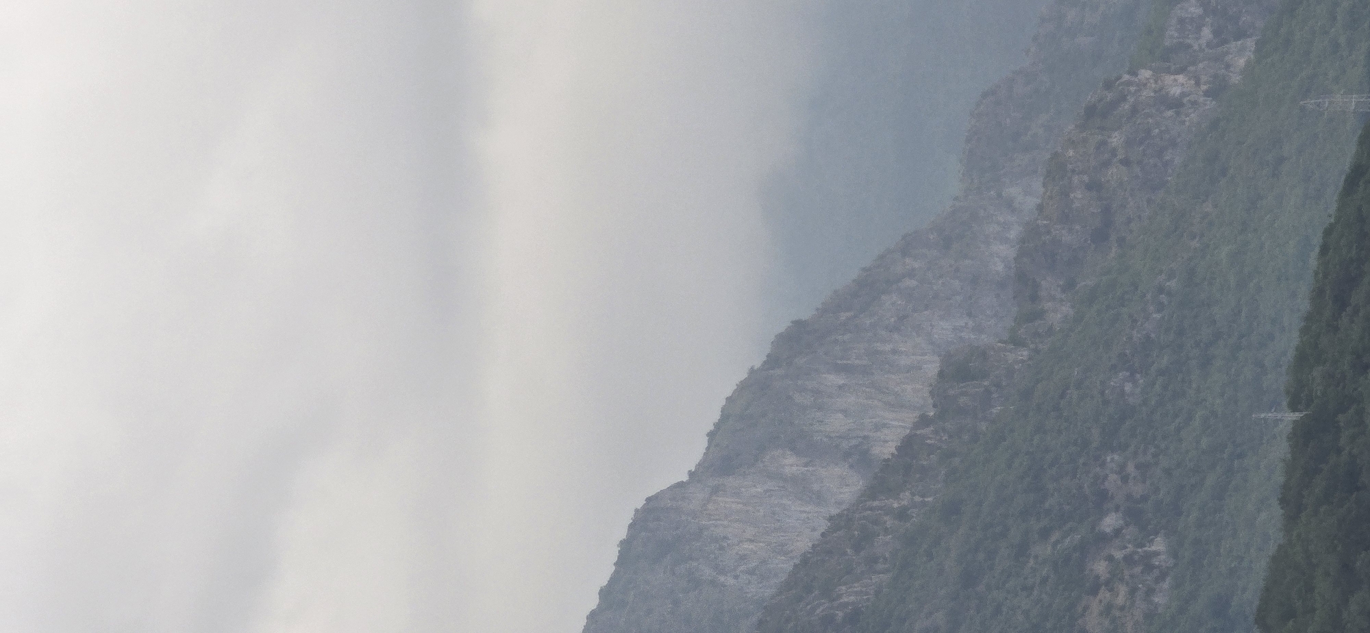 The image captures a hazy, overcast mountainous landscape. The sky, filling the upper left portion, is a uniform light gray, indicating heavy cloud cover with no direct sunlight.
The scene features multiple layers of mountains receding into the distance. The background mountains appear muted and bluish due to atmospheric haze, while the closer ranges show more detail. These midground and foreground mountains are characterized by a mix of rugged, exposed rock faces and dense green vegetation, primarily trees. Large sections of the mountains reveal grayish, rocky surfaces, interspersed with dark green, forested areas that cling to the slopes. The outlines of the mountain peaks and ridges are softened by the diffused light and haze.
Faint, horizontal linear structures, possibly elements of power lines or some form of infrastructure, are subtly visible traversing the forested slopes towards the right side of the image. No people or discernible activities are present. The lighting suggests an overcast day, contributing to the serene and somewhat subdued mood of the natural setting. There is no visible text. The image offers a broad view of a rugged, natural environment, consistent with mountainous regions such as Bar, Montenegro.