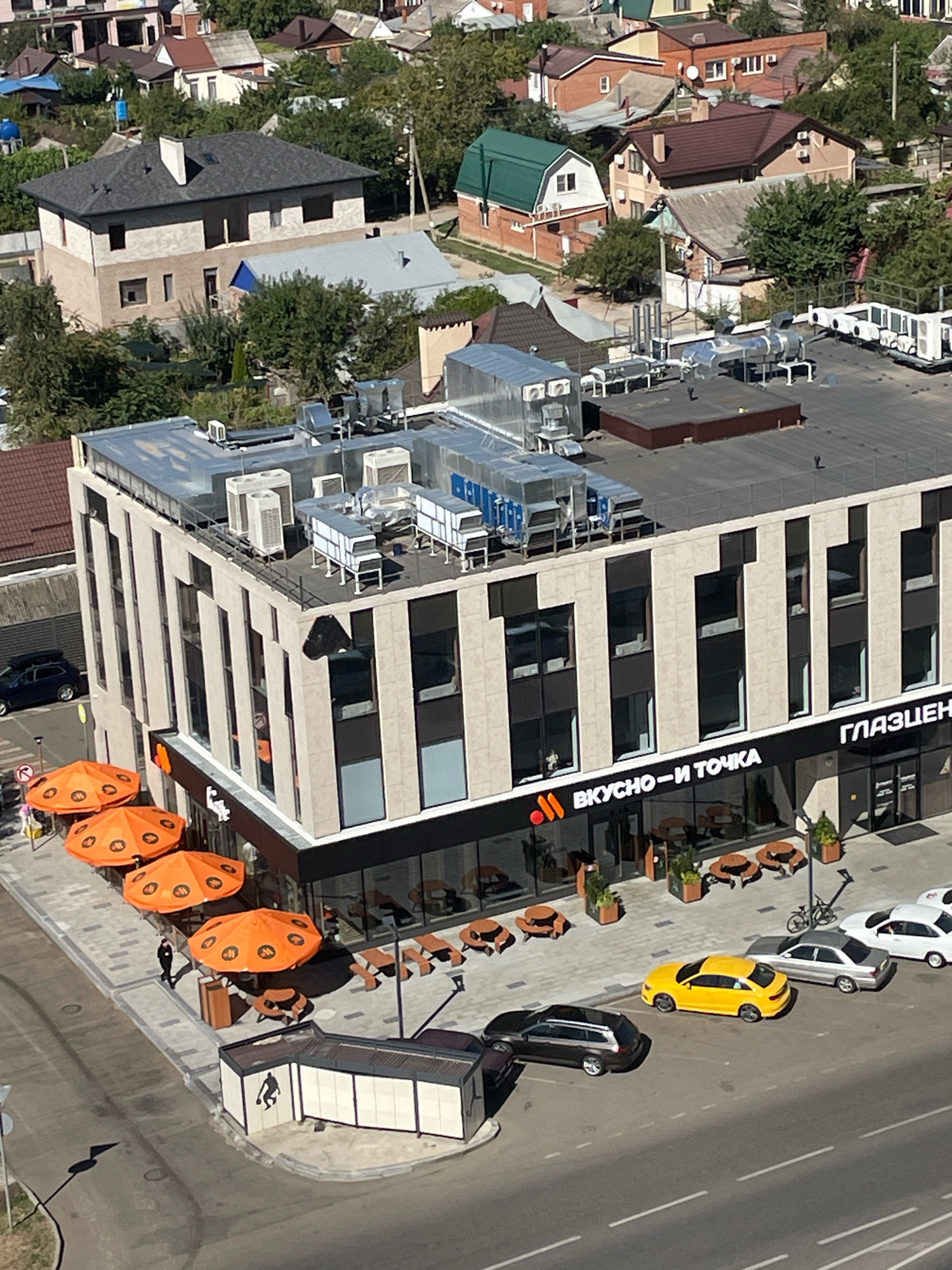 An aerial view captures a modern commercial building in the city of Krasnodar, Russia, with a bright blue sky overhead. The building features a facade of light-colored vertical panels and large dark windows. On its rooftop, several air conditioning units and ventilation systems are visible. The ground level of the building houses a restaurant with an outdoor seating area, identifiable by orange umbrellas bearing a logo and picnic-style tables. Adjacent to the restaurant, a sign displays the text "ВКУСНО-И ТОЧКА" in Russian, which translates to "Tasty - and that's it."
In front of the building, on a paved walkway, are several outdoor seating arrangements covered by orange umbrellas. A person in dark clothing is walking near the seating area. Further down the street, a parking area is occupied by several cars, including a yellow sedan, a dark-colored station wagon, a silver sedan, and a white sedan. A bicycle is parked near the building. The surrounding area is characterized by residential buildings with red and beige roofs, interspersed with lush green trees, suggesting a suburban or mixed-use urban environment.
