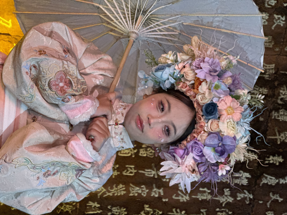 A young individual, likely female, is centered in a medium shot, facing slightly right with a neutral expression. The subject wears a traditional-style, pale beige or light pink jacket with embroidered patterns and pink fur trim on the voluminous cuffs. A standing collar is secured by a red-jeweled clasp. An elaborate floral headdress, composed of numerous artificial flowers in shades of pink, purple, blue, white, and green, adorns the head. Facial makeup includes prominent eyeliner and subtle glitter or decorative elements on the cheeks. The subject holds a light-colored traditional paper umbrella with a wooden handle resting on their left shoulder. The right hand is positioned near the chest. The background is a dark, textured surface, possibly stone, with numerous illuminated Chinese characters carved or painted, indicating a cultural or historical location within Yuzhong District, China.