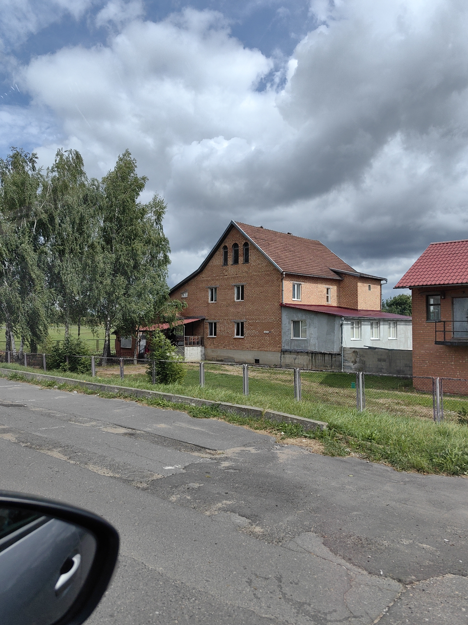 In Myadzyel, Belarus, a side-view mirror of a vehicle is visible in the foreground, reflecting a cloudy sky. The road ahead shows signs of wear and tear, with patches and cracks. Beyond the road, a grassy area is bordered by a fence. Behind the fence, two brick buildings with red tiled roofs are present. The larger building on the left is a two-story structure with several windows and an attic. A smaller, single-story extension with grey walls and white-framed windows is attached to its side. To the right, another brick building with a red roof features a window and a balcony. Several tall trees with green foliage are situated between the road and the buildings.