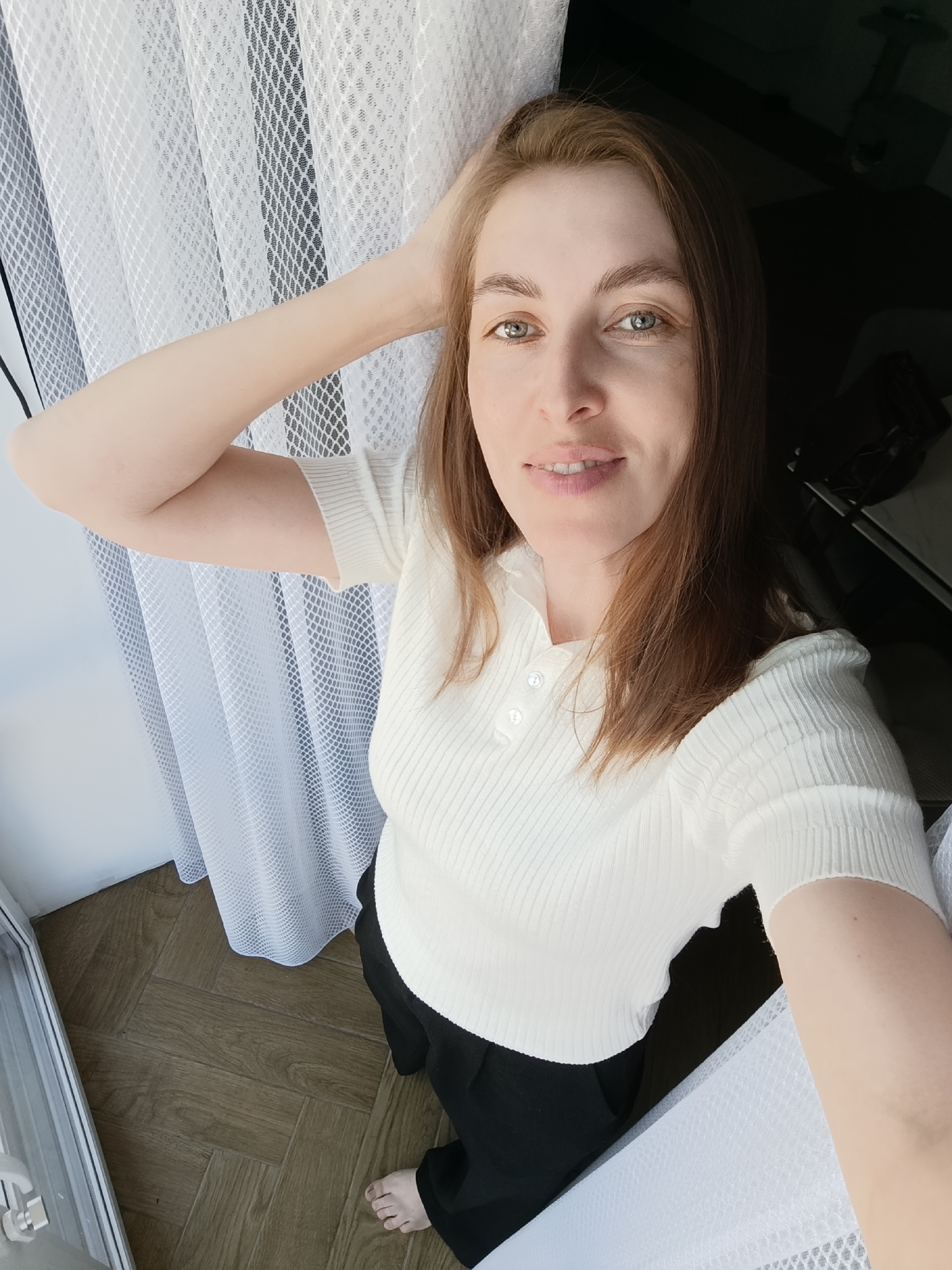 A medium shot of a light-skinned woman indoors near a window. She wears a white, short-sleeved, ribbed shirt with buttons at the neck and black pants. Her hair is light brown, and she is holding one hand behind her head. The window has a white, semi-transparent curtain. The floor is covered with wooden planks arranged in a herringbone pattern. Part of a bare foot is visible in the bottom left corner. The location appears to be a room in a house or apartment in Chaikovsky, Russia.