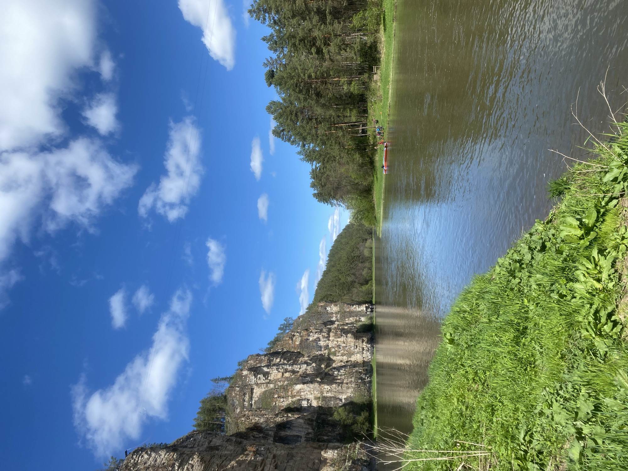 A landscape photograph taken in Kul'metovo, Russia. The image features a river running between a rocky cliff on the left and a grassy bank on the right. A line of trees sits atop the cliff. The sky above is blue with scattered white clouds. A red boat is visible in the distance on the river. The image is oriented with the top of the scene to the left.
